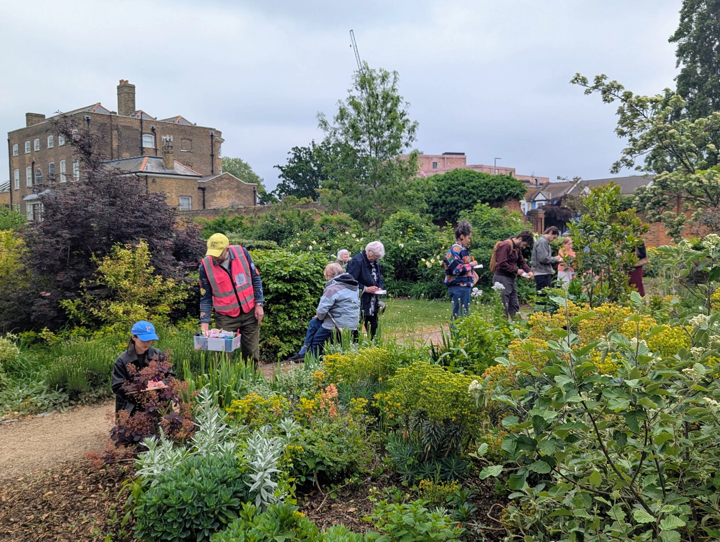 People gardening in a lush green garden with various plants, trees, and blooming flowers, with residential buildings and a crane in the background on a cloudy day.