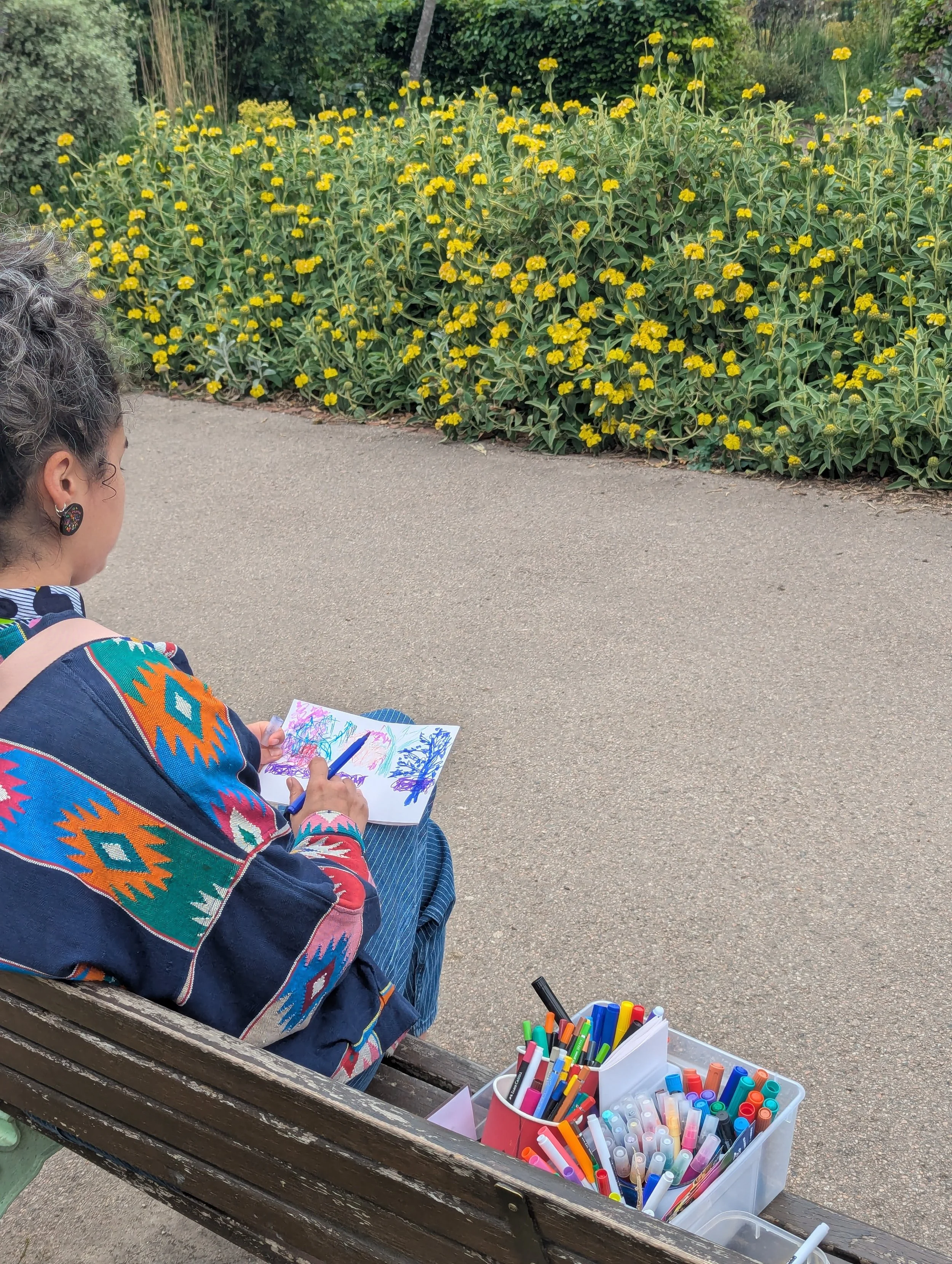 A woman sits on a park bench with colorful markers and drawings, facing yellow flowering plants and greenery.