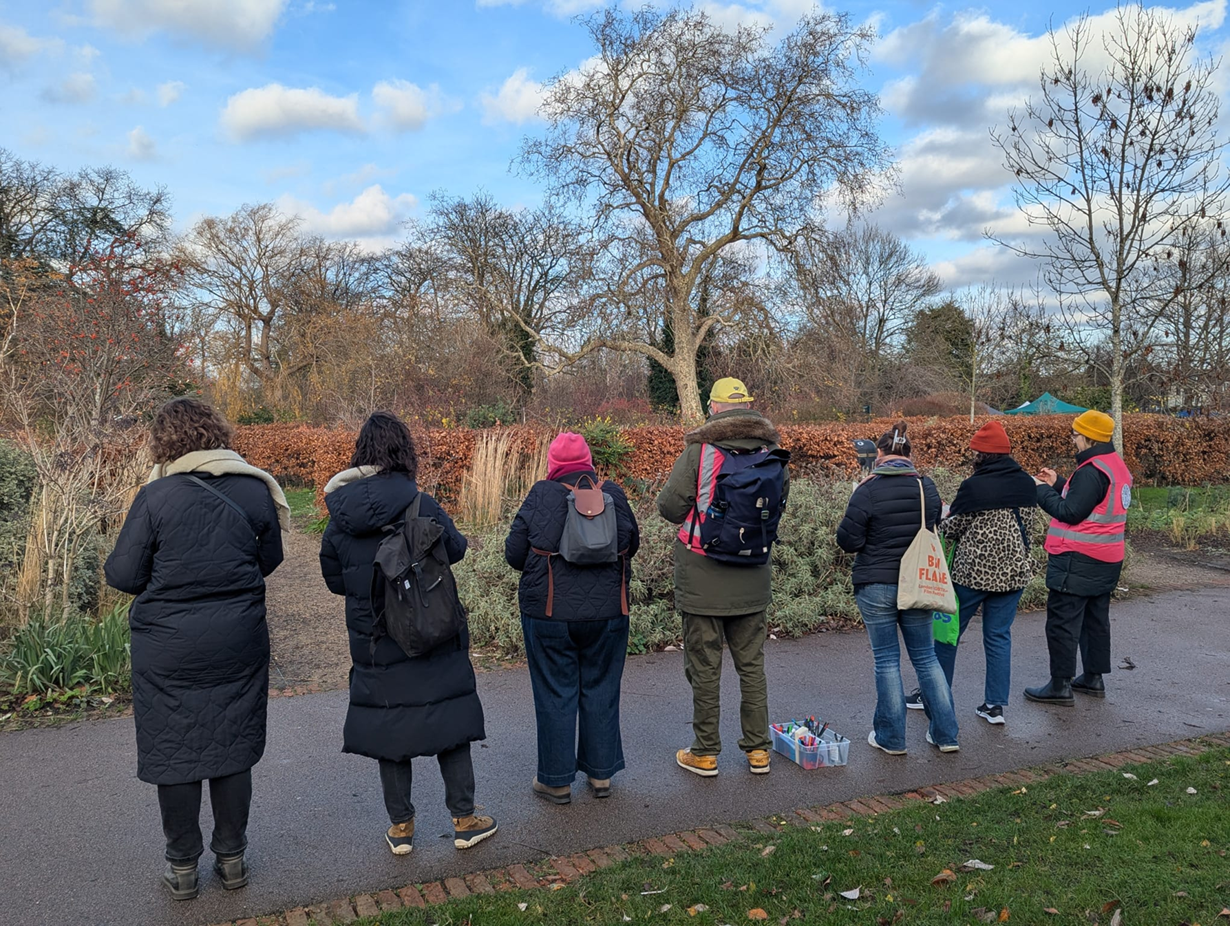 Group of people standing outdoors in a park during autumn, facing away from the camera, with bare trees and a blue sky with scattered clouds in the background.
