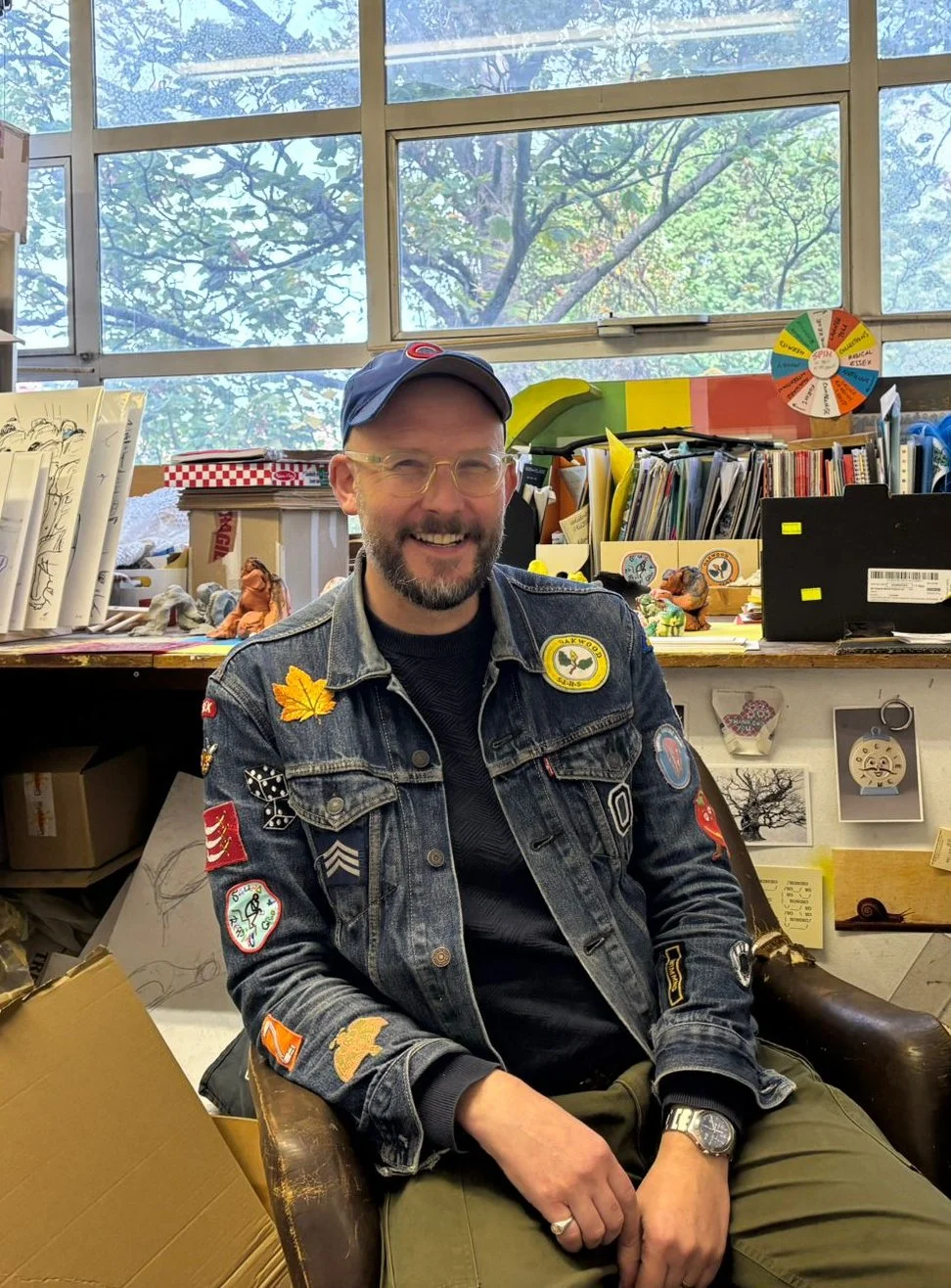 Barry Sykes with glasses, a beard, and wearing a blue baseball cap and a denim jacket with patches and embroidered patches, sitting in a cluttered workspace with books, papers, and decorations, in front of a large window showing trees outside.