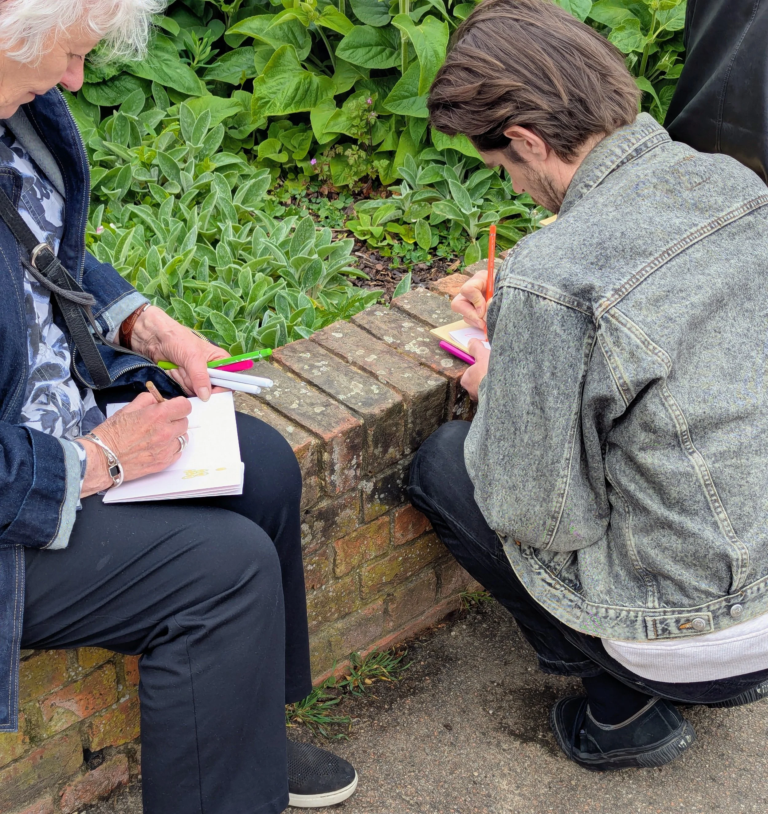 Three people are outdoors near a brick wall, leaning over a written or drawn document on a piece of paper. Only parts of their bodies are visible: a woman with white hair on the left, a man with brown hair wearing a gray denim jacket on the right, and a person in black on the far right. The background features lush green plants.