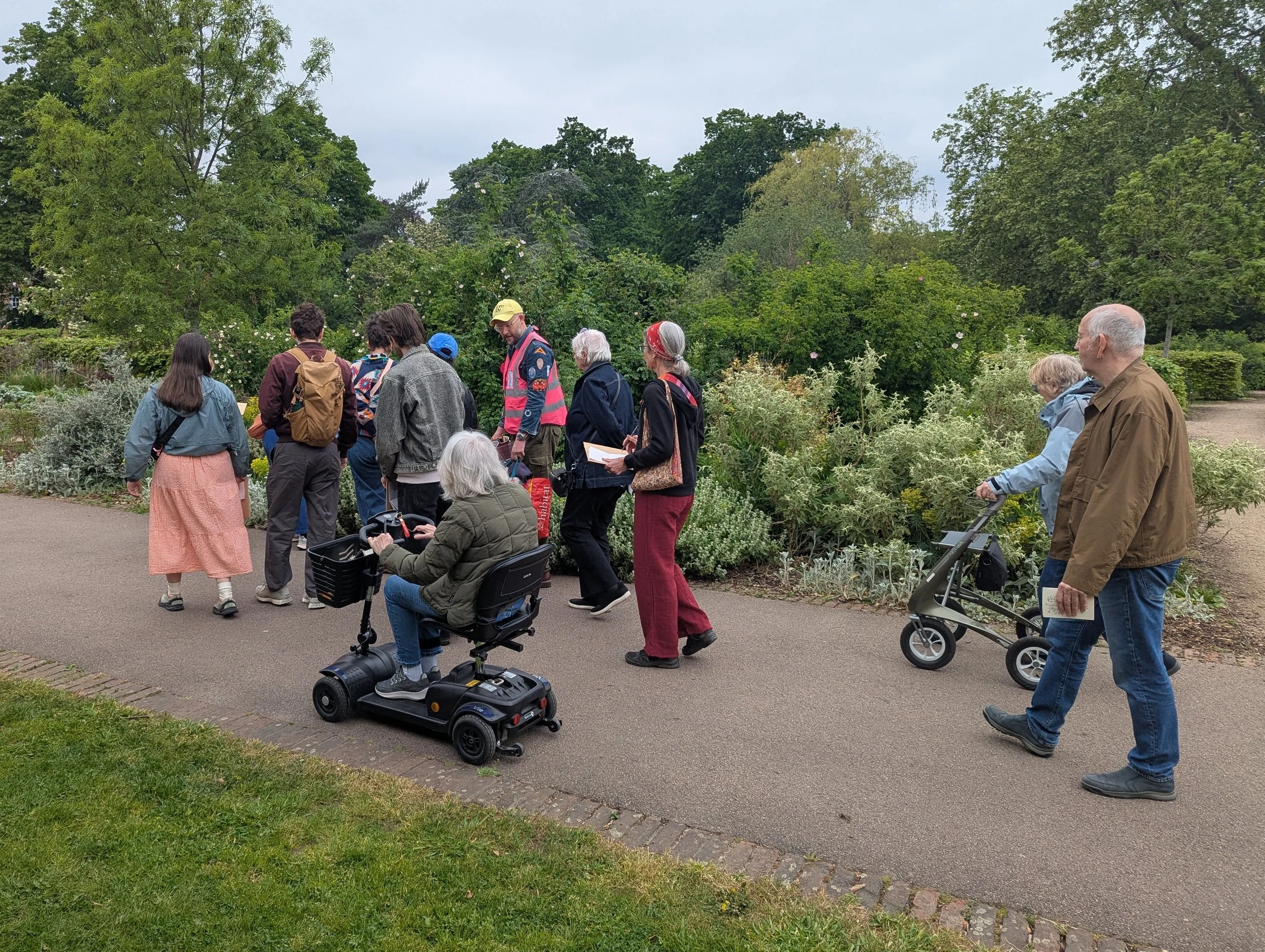 A group of people gathered outdoors in a park-like setting with trees and greenery in the background. Some are standing, others are sitting or using mobility devices, and they appear to be participating in a walk or outdoor event.