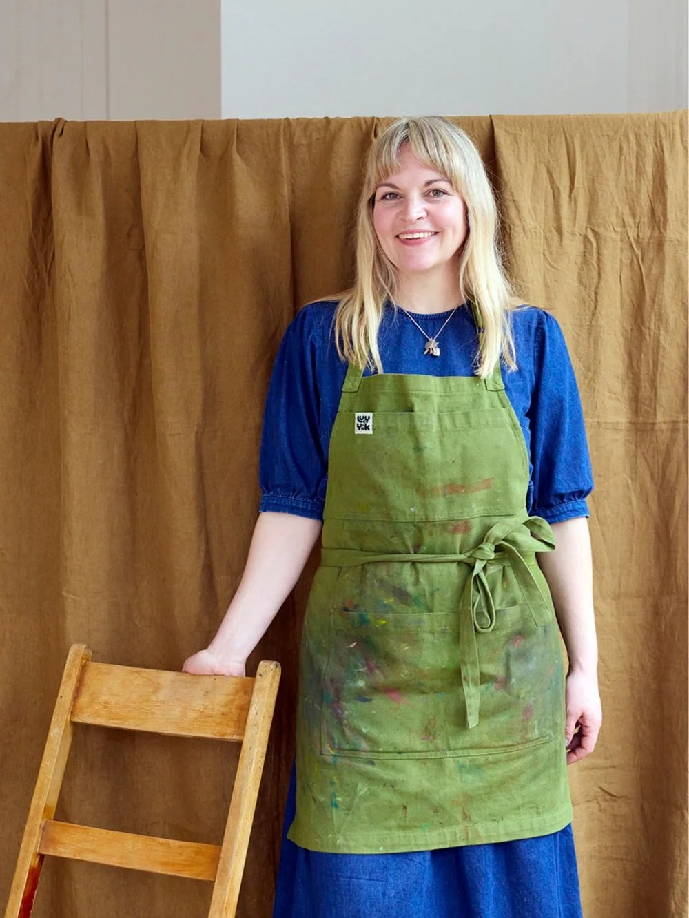 A woman artist, Emma Bond with blonde hair, smiling, wearing a blue dress and a green aprons with paint stains, standing in front of a brown curtain, holding a wooden chair.