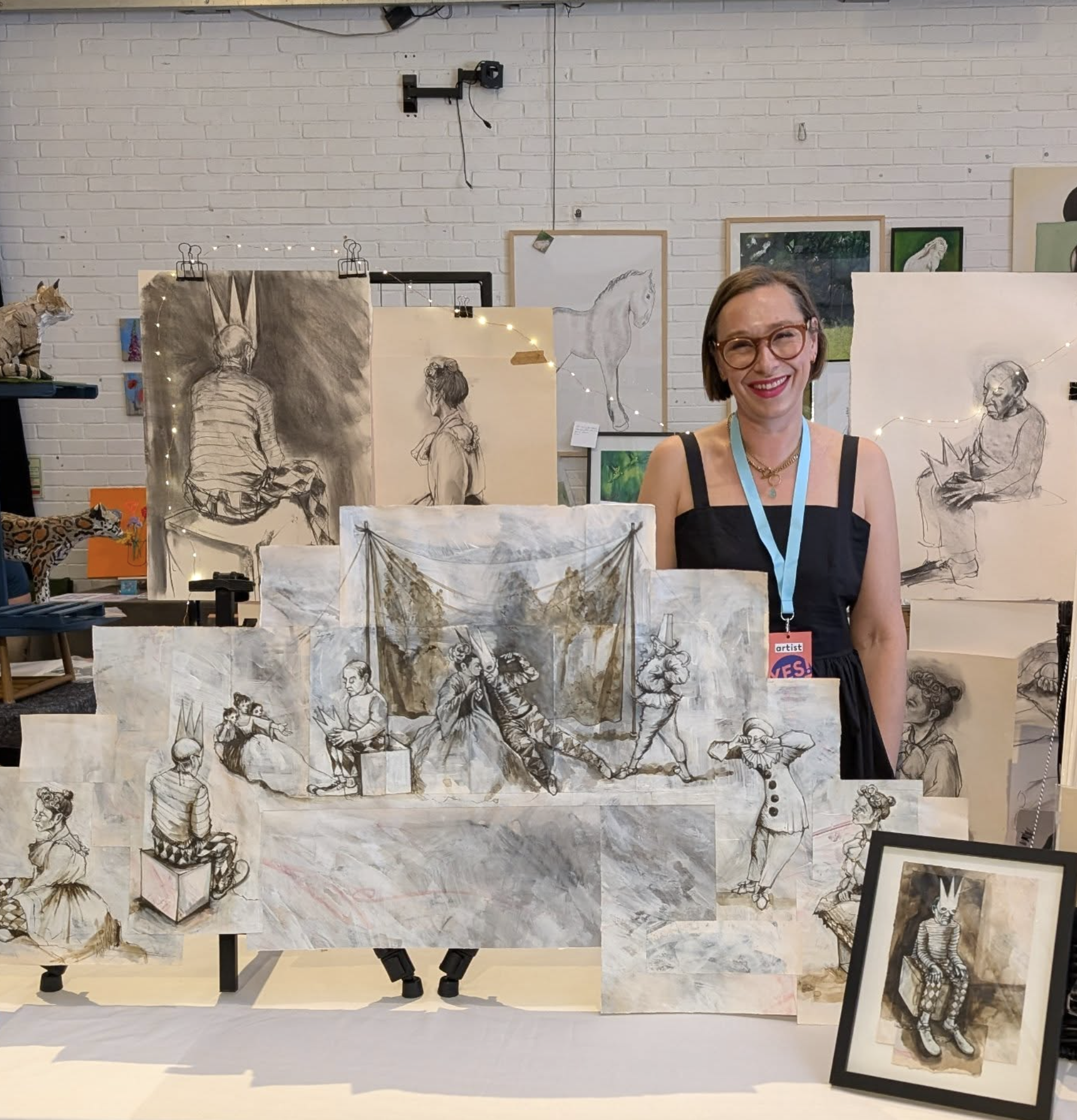 Ellie Horry at her art exhibition displays various sketches and drawings of people and animals, including a woman with glasses smiling in the foreground, and artwork of a horse, a tiger, and a seated person, with a white brick wall in the background.