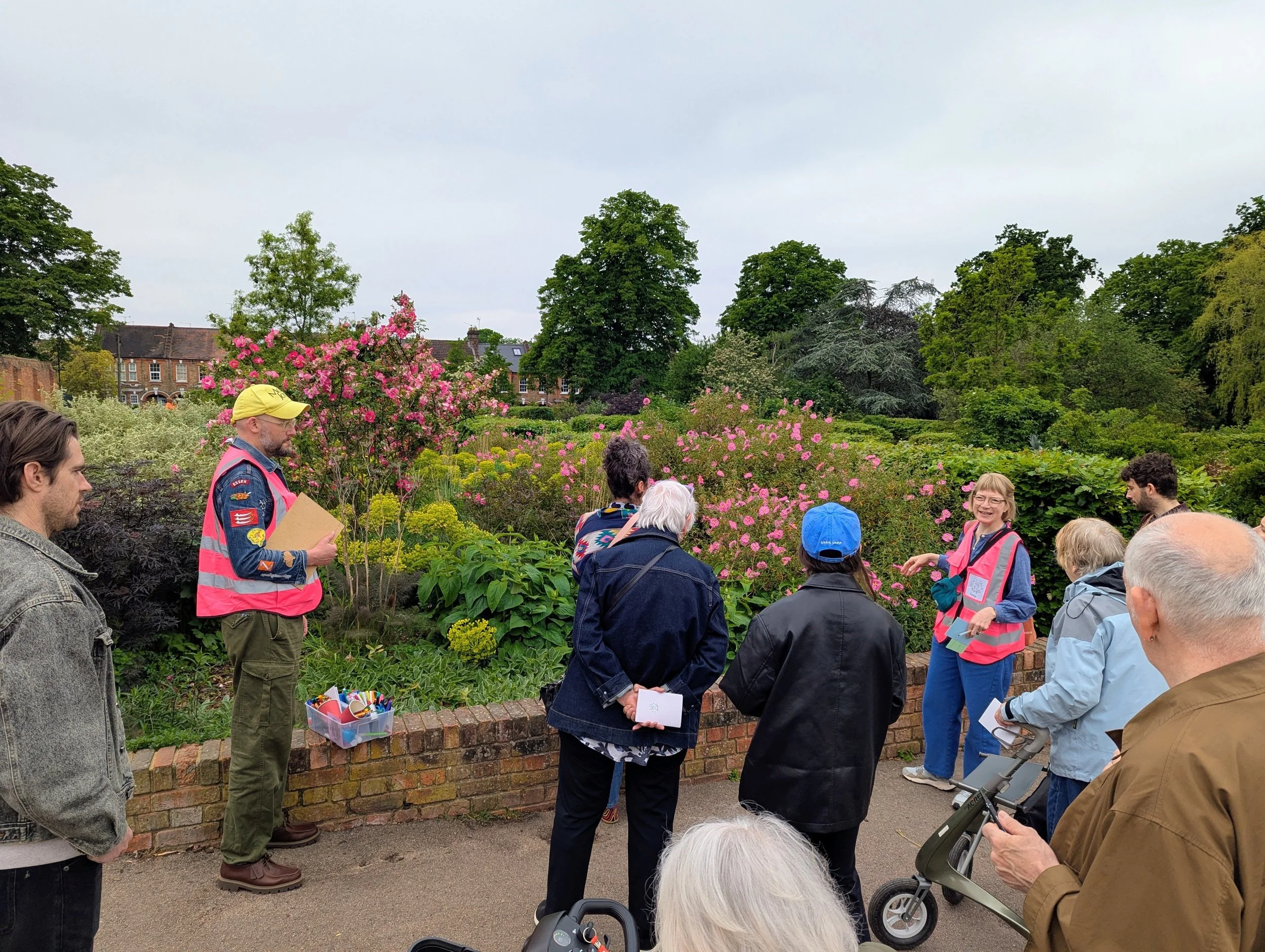 A group of people gathered outdoors in a park with lush green trees and pink flowering bushes, some holding papers or electronic devices, with a person wearing a yellow cap and pink vest appearing to speak or present.