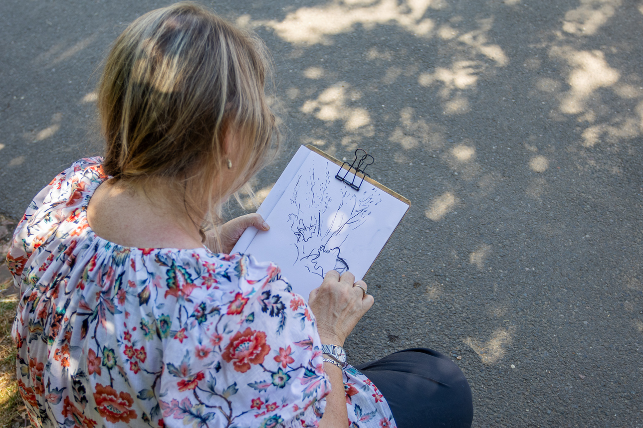 A woman with light brown hair wearing a floral shirt is sitting on the ground, drawing on a clipboard with a black clip. She is sketching trees and a bird outdoors on a paved path with shadows of leaves.
