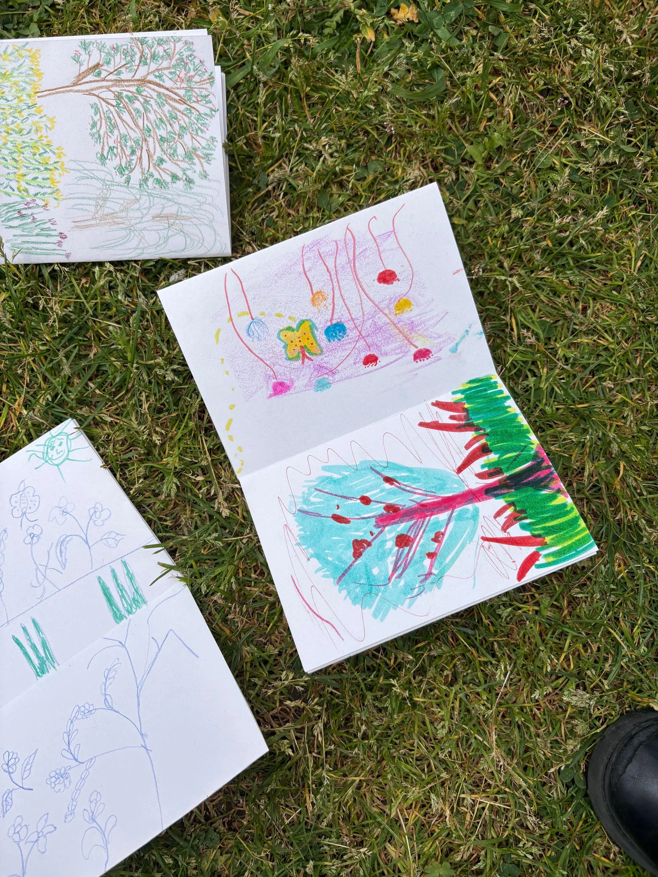 Colorful children's drawings of trees, flowers, and abstract shapes are scattered on green grass.