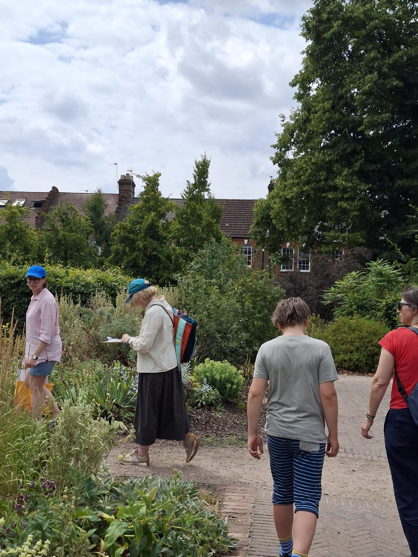 People walking in a lush garden with various plants and flowers, some looking at notes or taking pictures, under a partly cloudy sky with trees and traditional brick houses in the background.