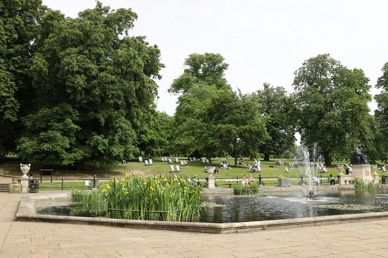 A park with a pond and fountain, surrounded by large trees and green grass. People are sitting on benches and lawn chairs, enjoying the day.