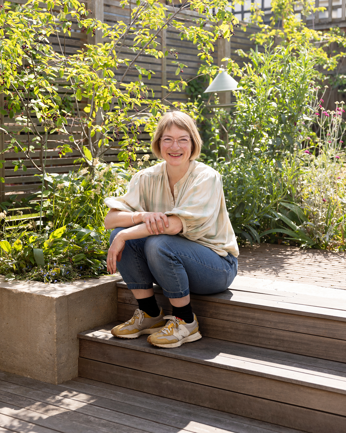 A woman with short blonde hair and glasses, smiling, sitting on wooden steps in a lush garden with green foliage and flowering plants.