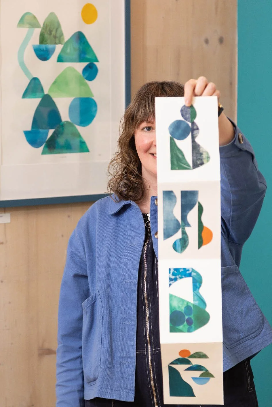 Siobhan Watt with curly brown hair wearing a blue jacket is holding a cutout with abstract shapes spelling out 'BROAD' in front of her face, with colorful abstract art on the wall behind her.