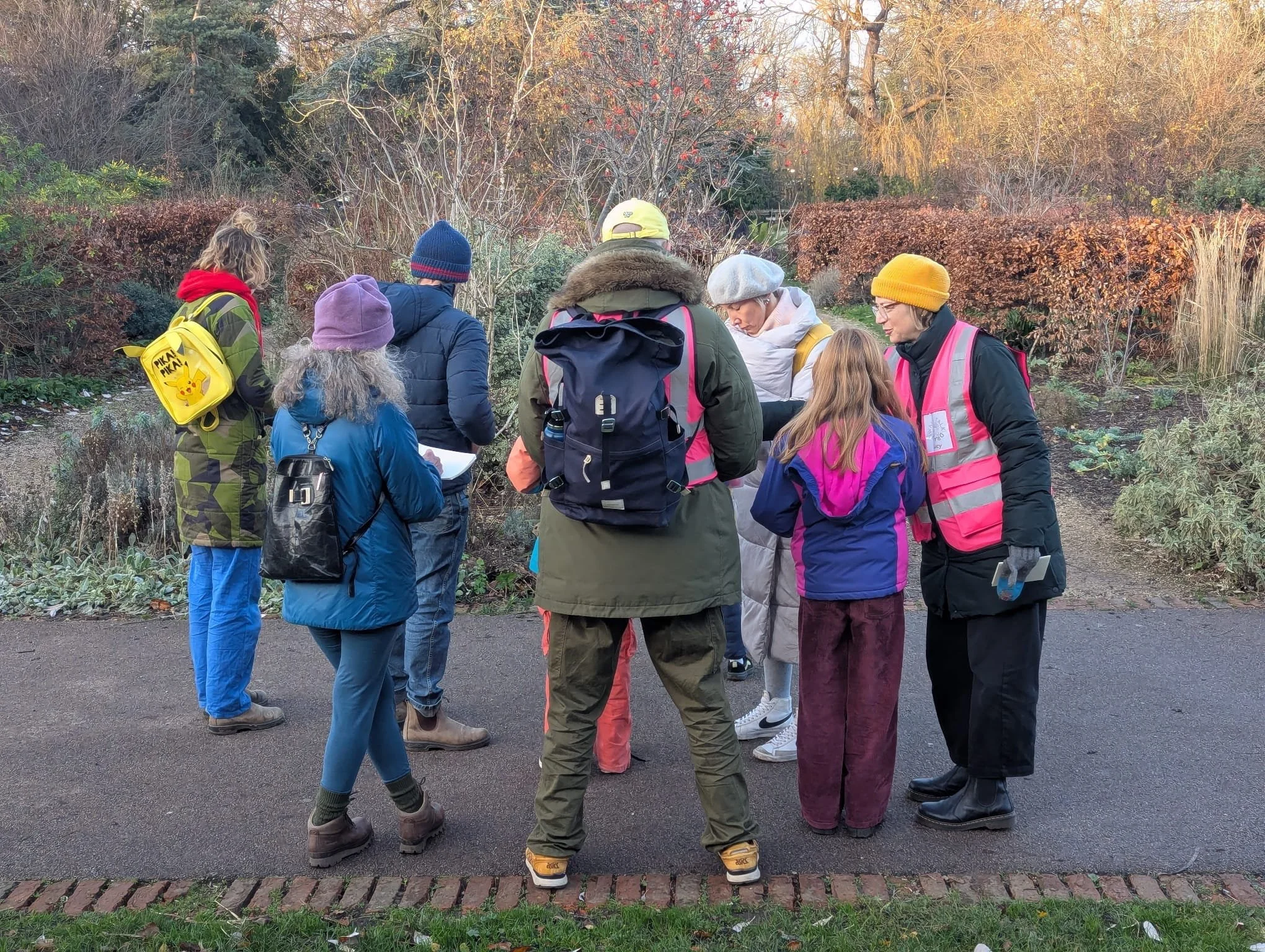 A group of people gathered outdoors, some wearing jackets and hats, in a garden with autumn foliage, listening to a woman with a white beret and glasses.