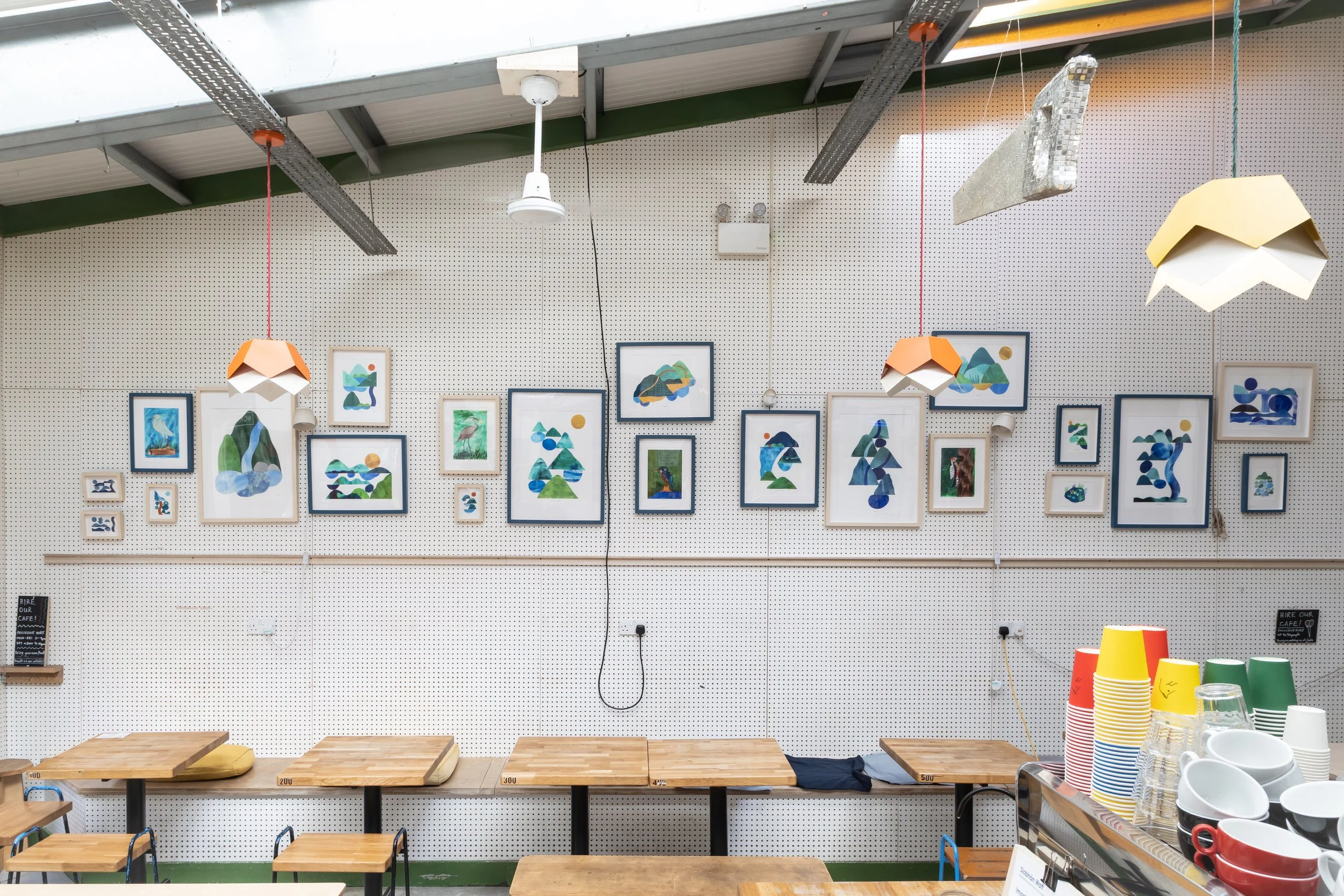 Interior of an art gallery or cafe featuring framed abstract artwork on a white pegboard wall, hanging pendant lights, and a display of colorful cups and bowls on a shelf.