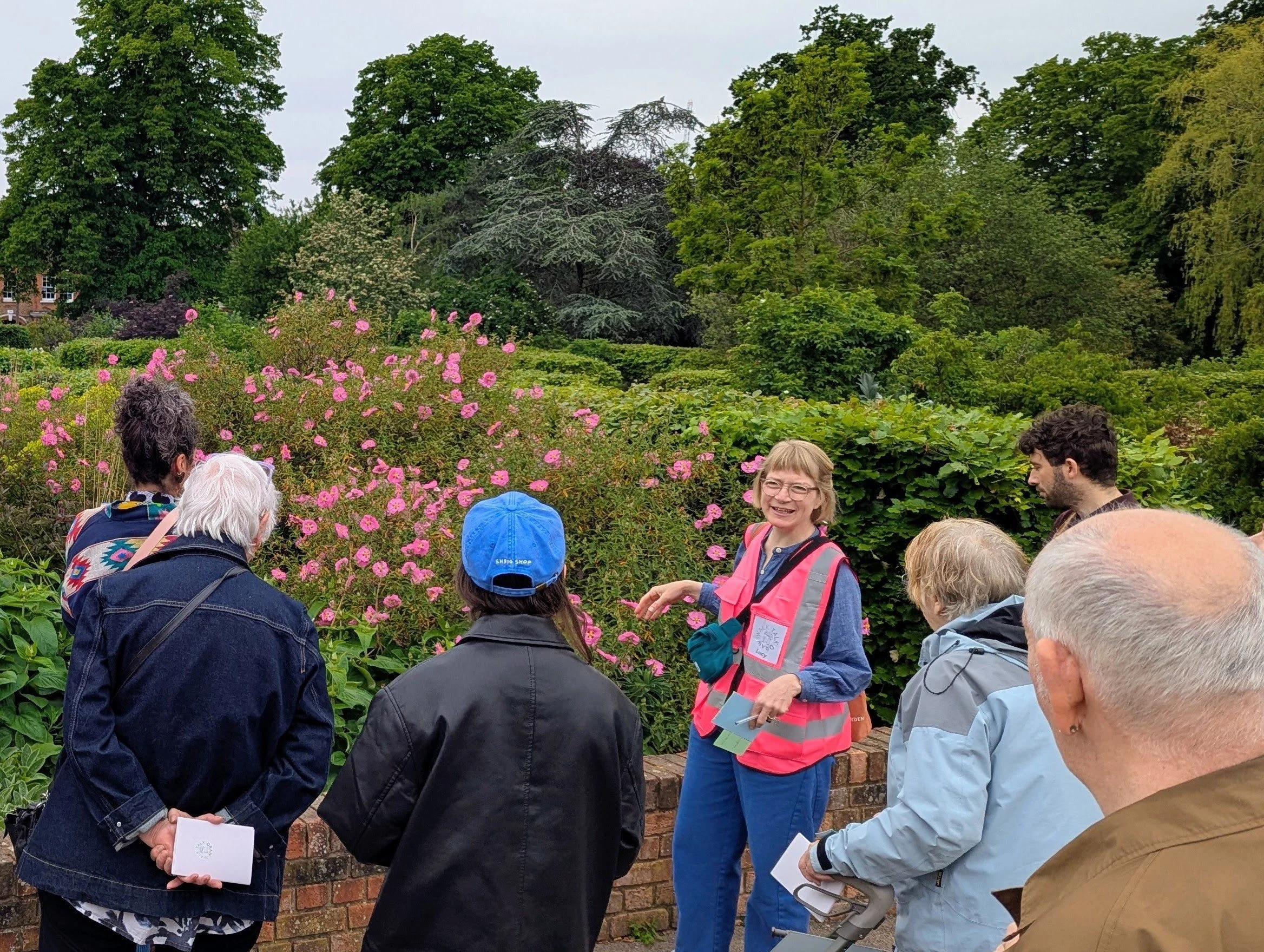 A group of people gathered in a garden with lush green trees and pink flowers, listening to a woman in a pink vest with a clipboard, who appears to be leading a tour or discussion.