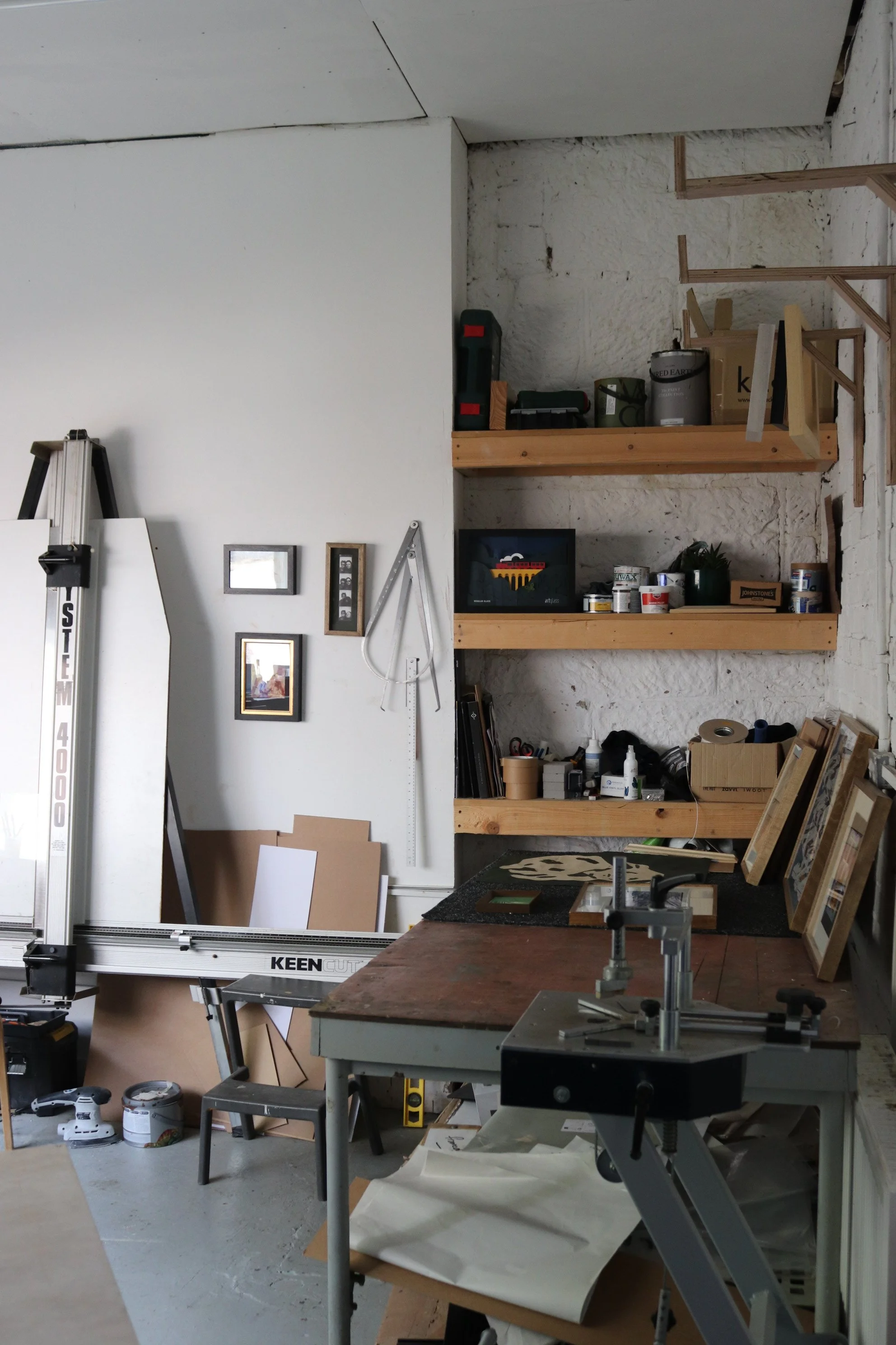 A lovely workshop with shelves holding tools and supplies, a worktable with a clamp, and various tools, materials, as well as framed pictures on the wall.