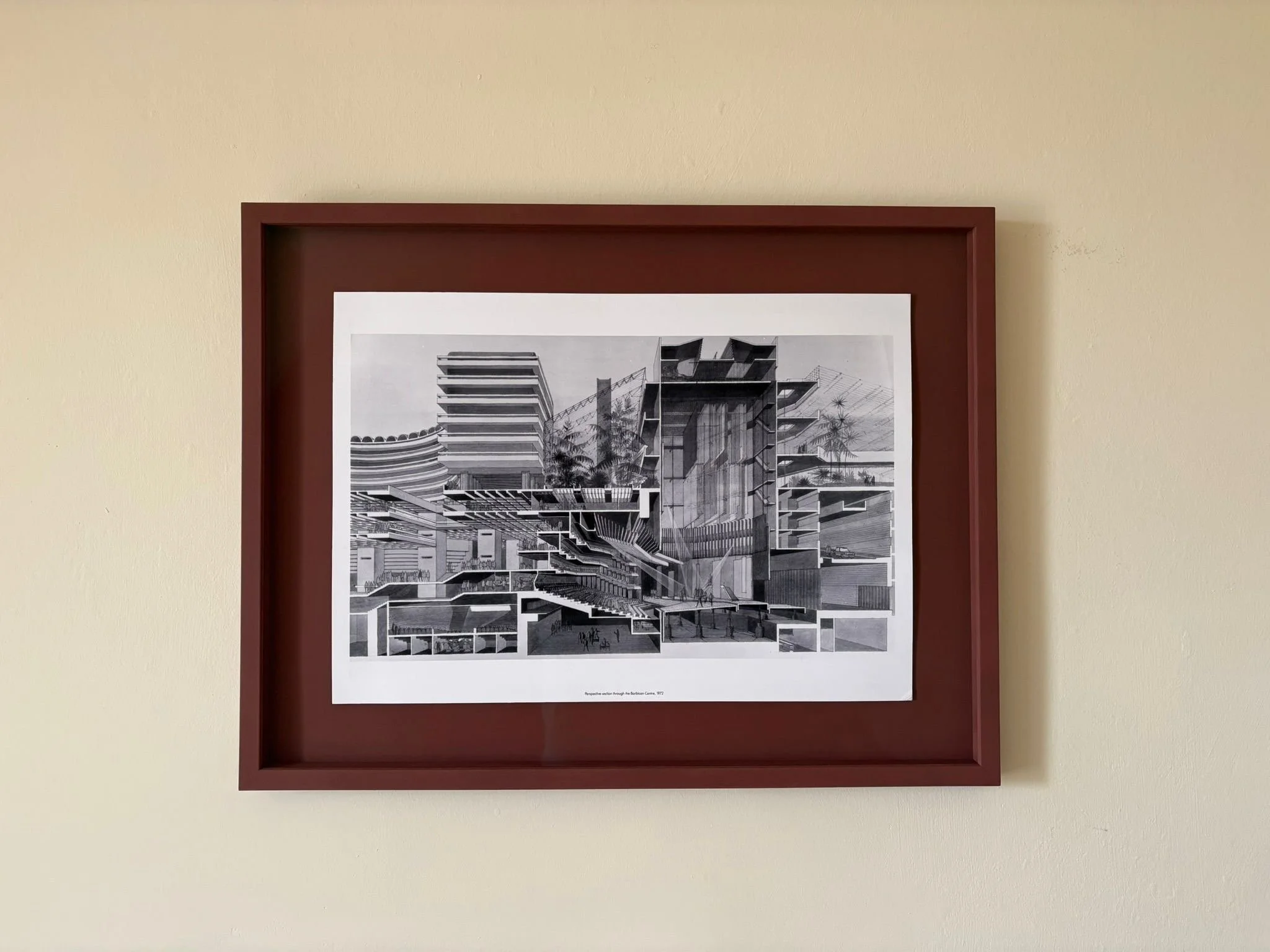 Black and white architectural drawing of a modern building with multiple levels, terraces, and palm trees, framed in reddish-brown, mounted on a beige wall.