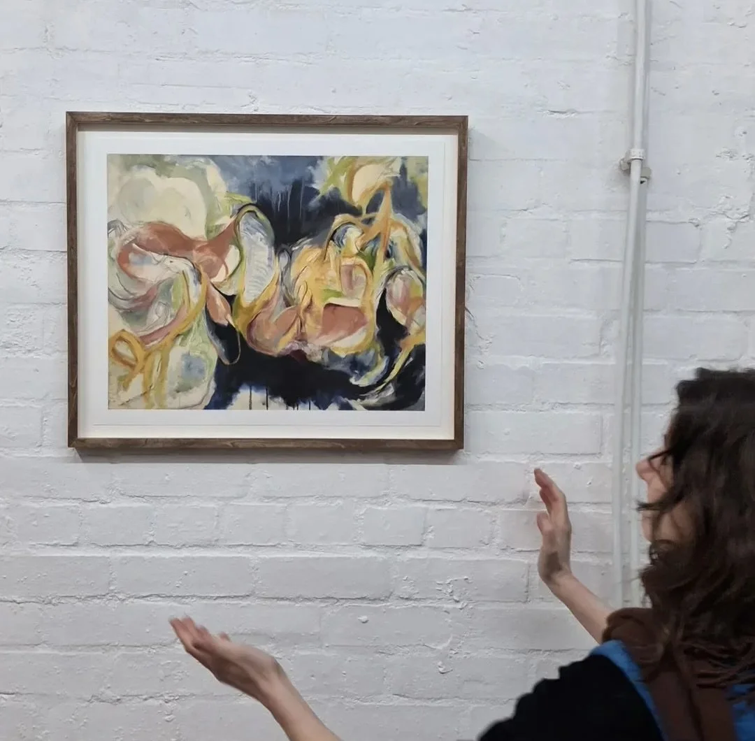 A woman in glasses gestures with her hands while viewing an abstract painting in an art gallery. The painting features swirling, colorful shapes on a white brick wall.