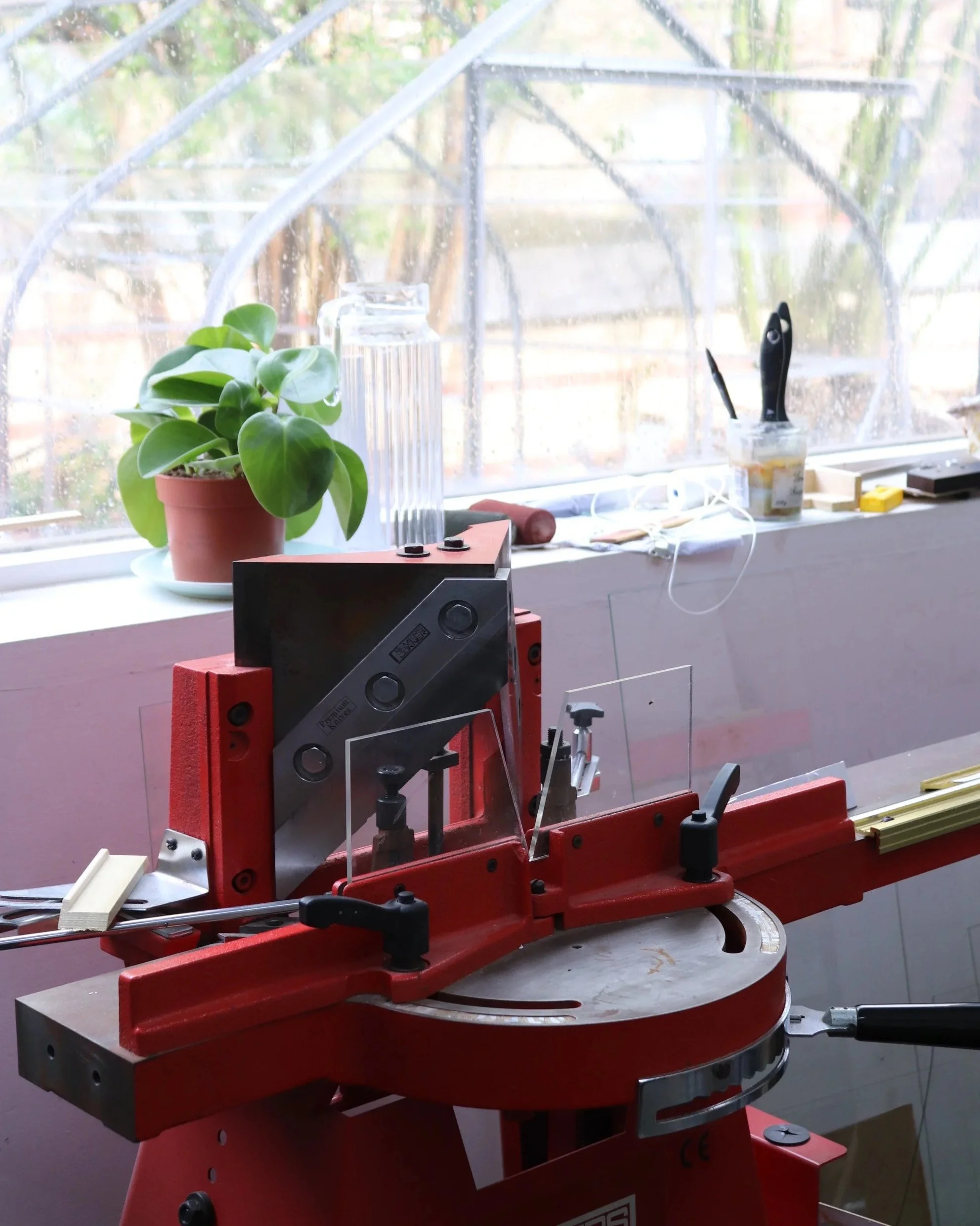 A red and white band saw with a machine saw blade, positioned in a workshop or studio, with a potted plant and various tools on the windowsill.