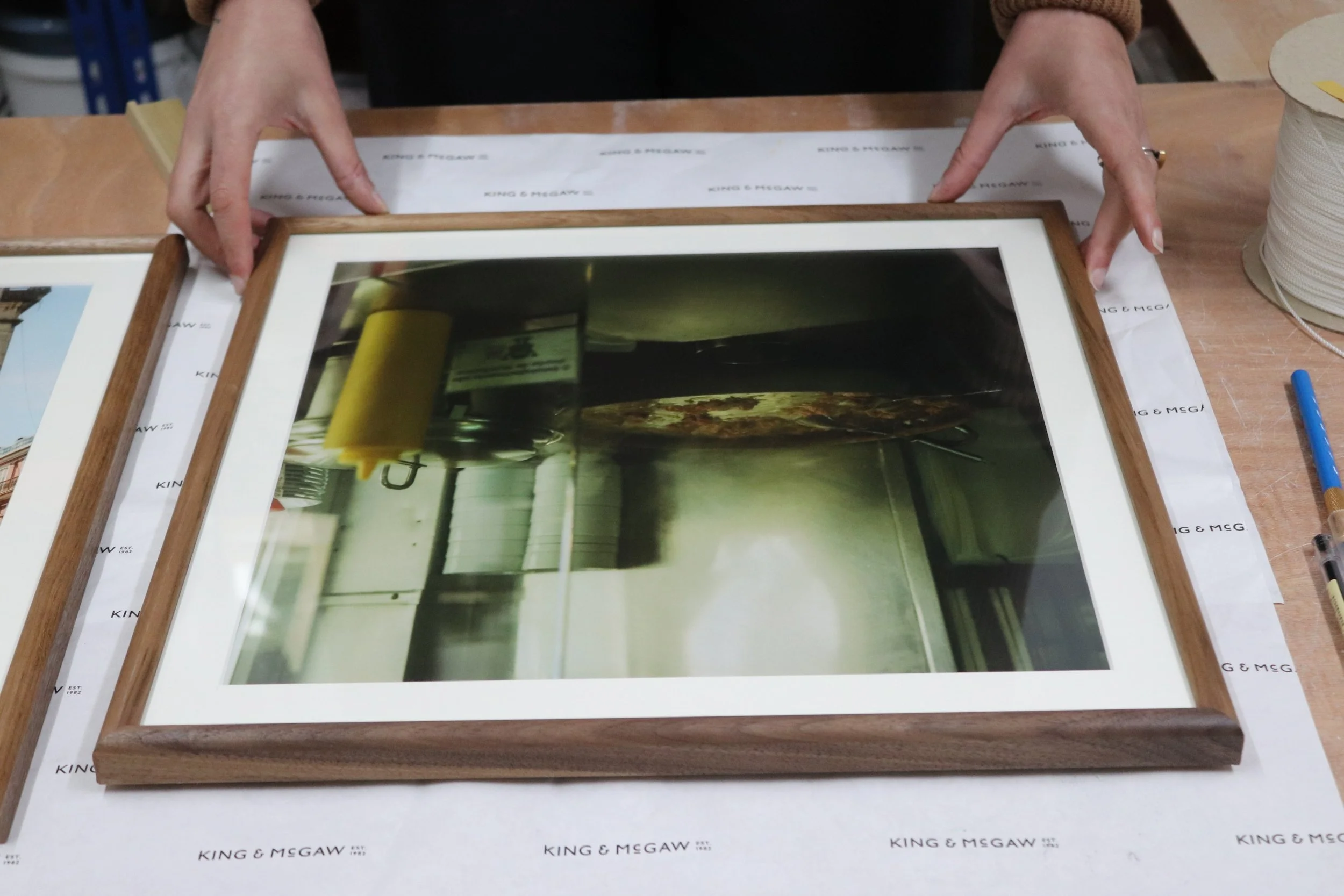 A person holding a beautiful framed photograph on a wooden table, with a roll of tape and a blue pen nearby.