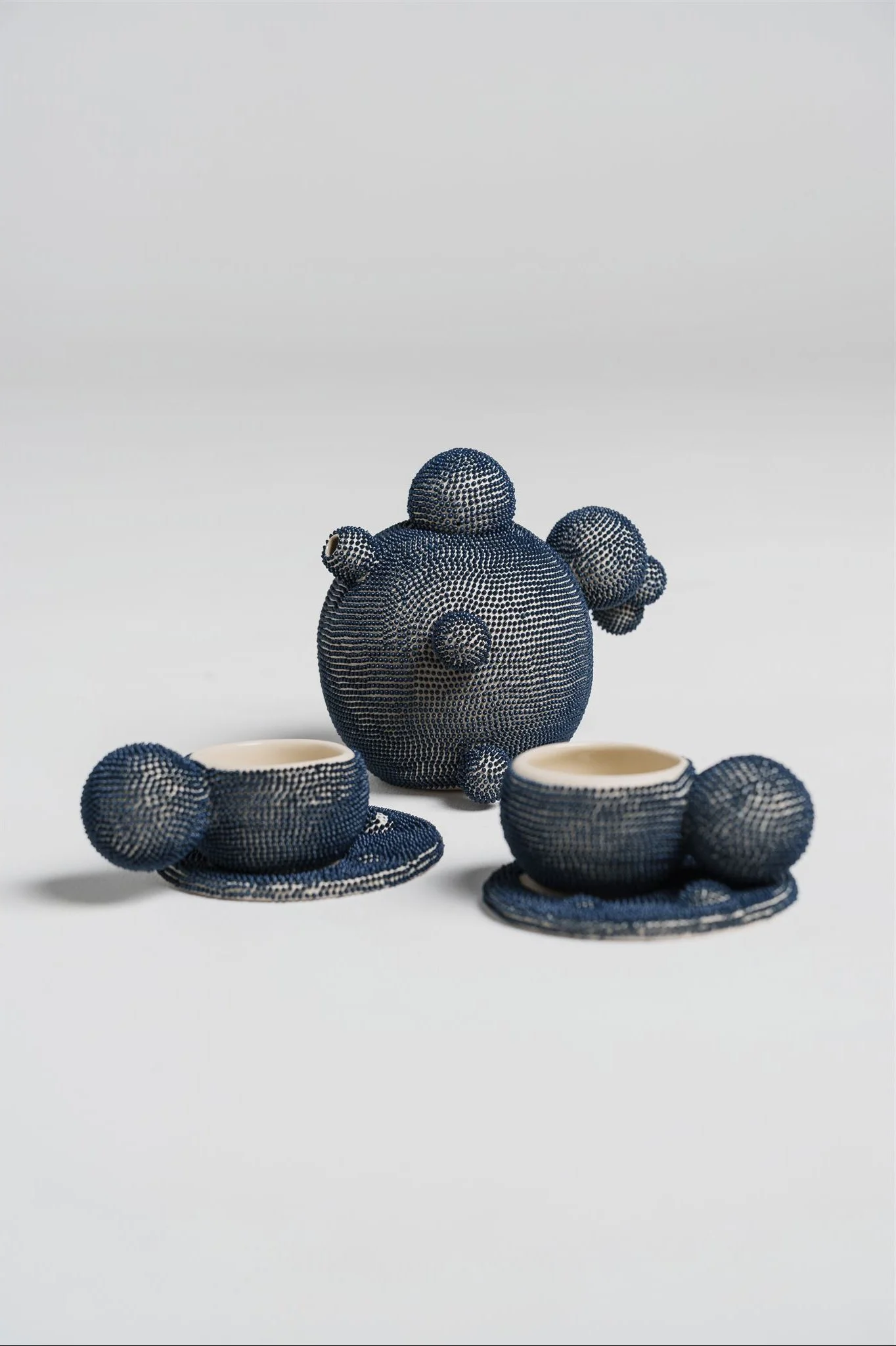 A three contemporary tea service featuring a teapot and two matching cups with saucers. Each piece is meticulously covered in a dense, uniform pattern of small indigo blue dots.