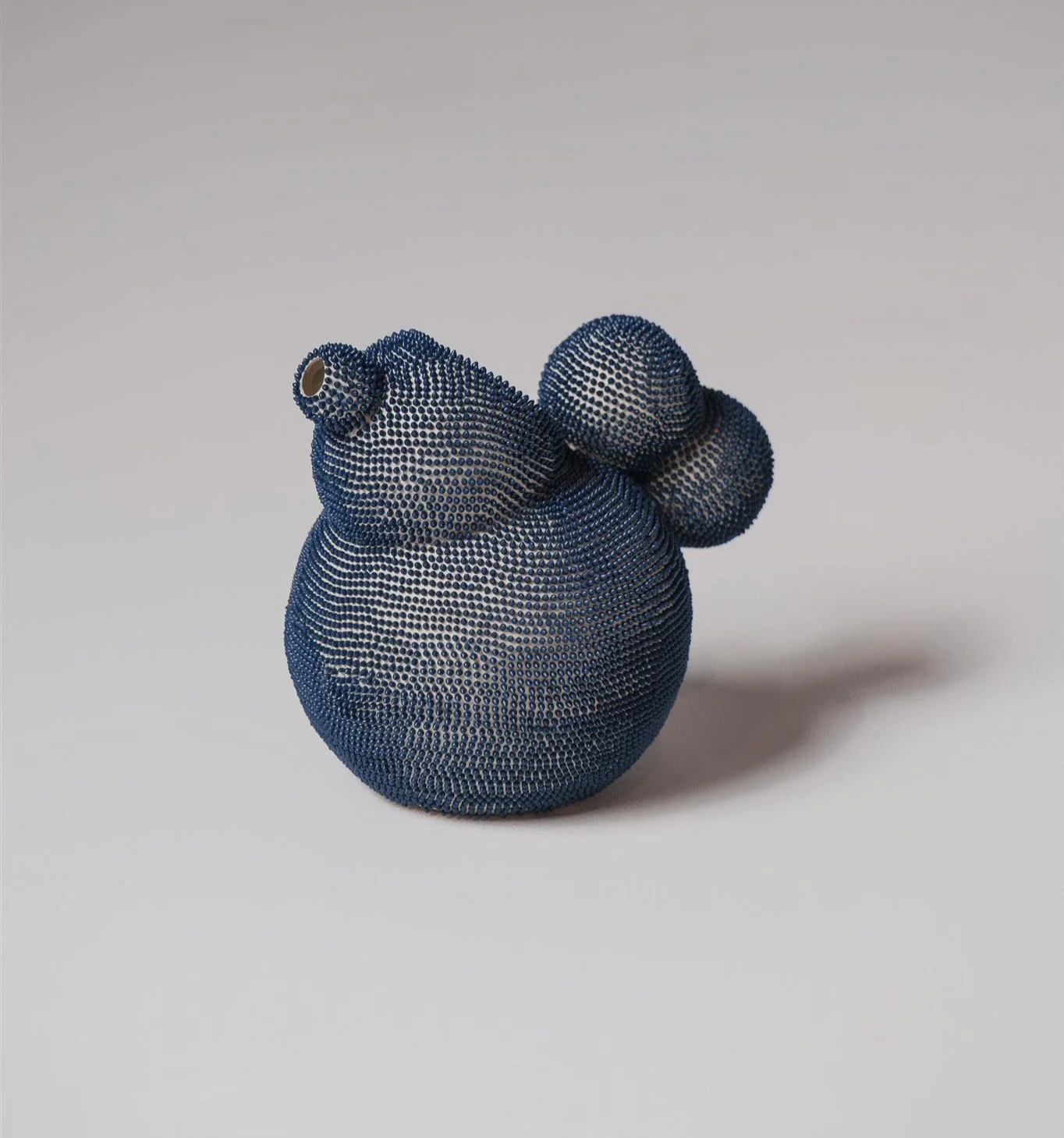 Studio photograph of a contemporary water jug with an organic, bulbous shape. The piece is made of smooth with ceramic and is covered in thousands of tiny, meticulously arranged navy-blue dots that create a dense, texture pattern over its surface.
