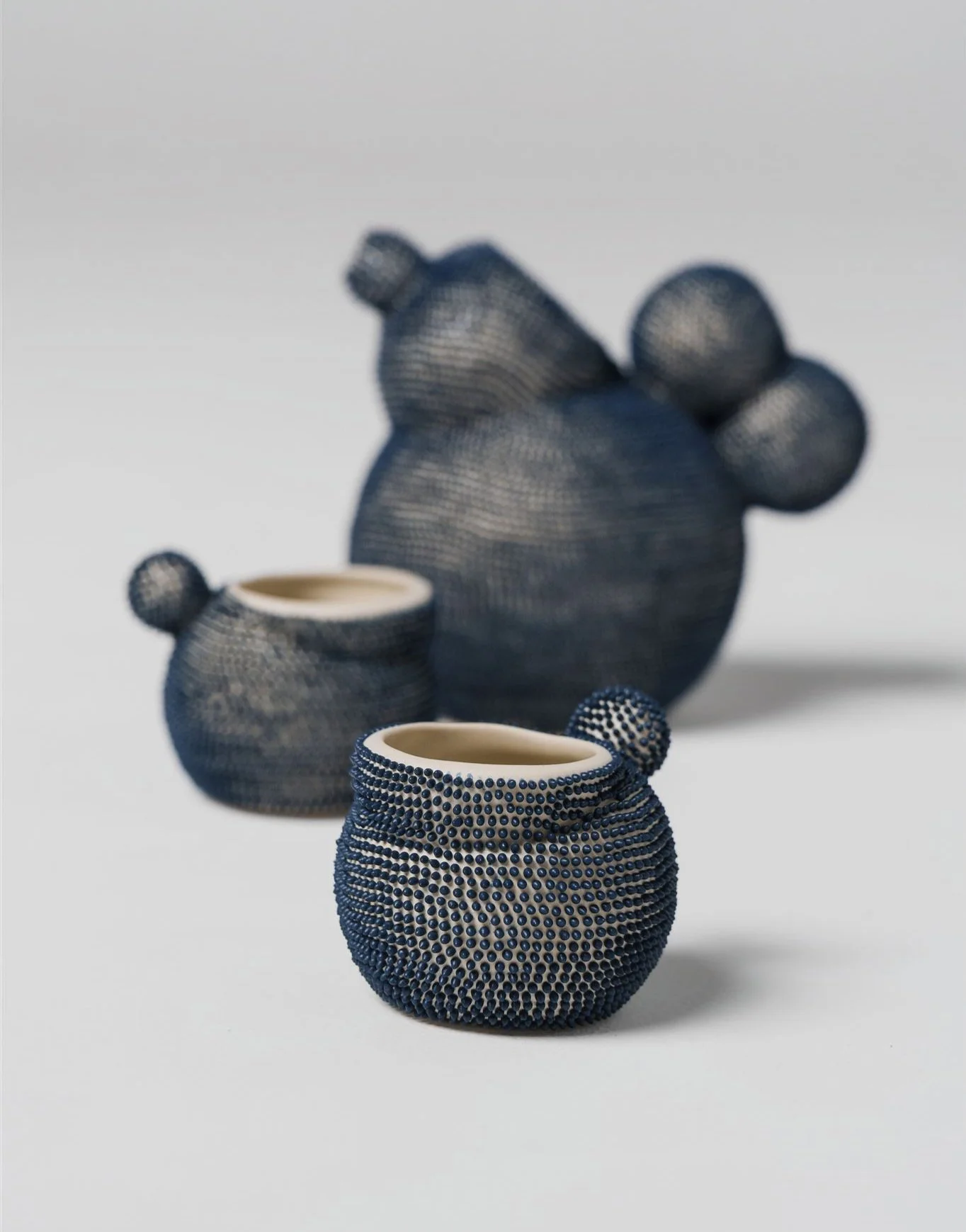 Studio photograph of three hand-built ceramic sculptures with rounded, organic forms, covered in dense blue engobe dots. The smallest glass in sharp focus in the foreground, with two other sculptural forms blurred in the background.