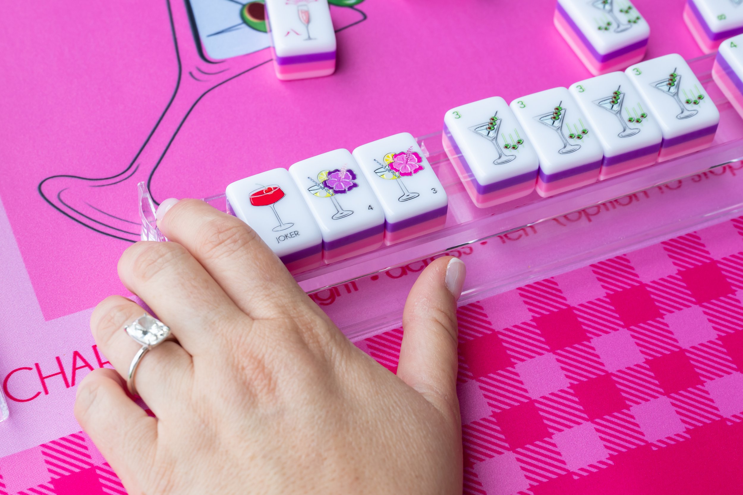 Close-up of a hand, holding mahjong tiles with tropical drink and martini illustrations, on a pink surface with a drawing of a cocktails and instructions for Charleston.