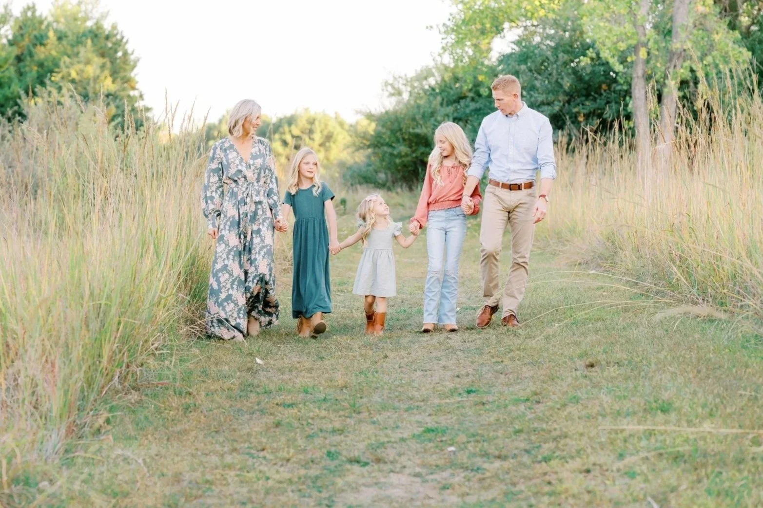 A family of six walking hand in hand down a grassy path surrounded by tall grass and trees, enjoying a sunny day outdoors.