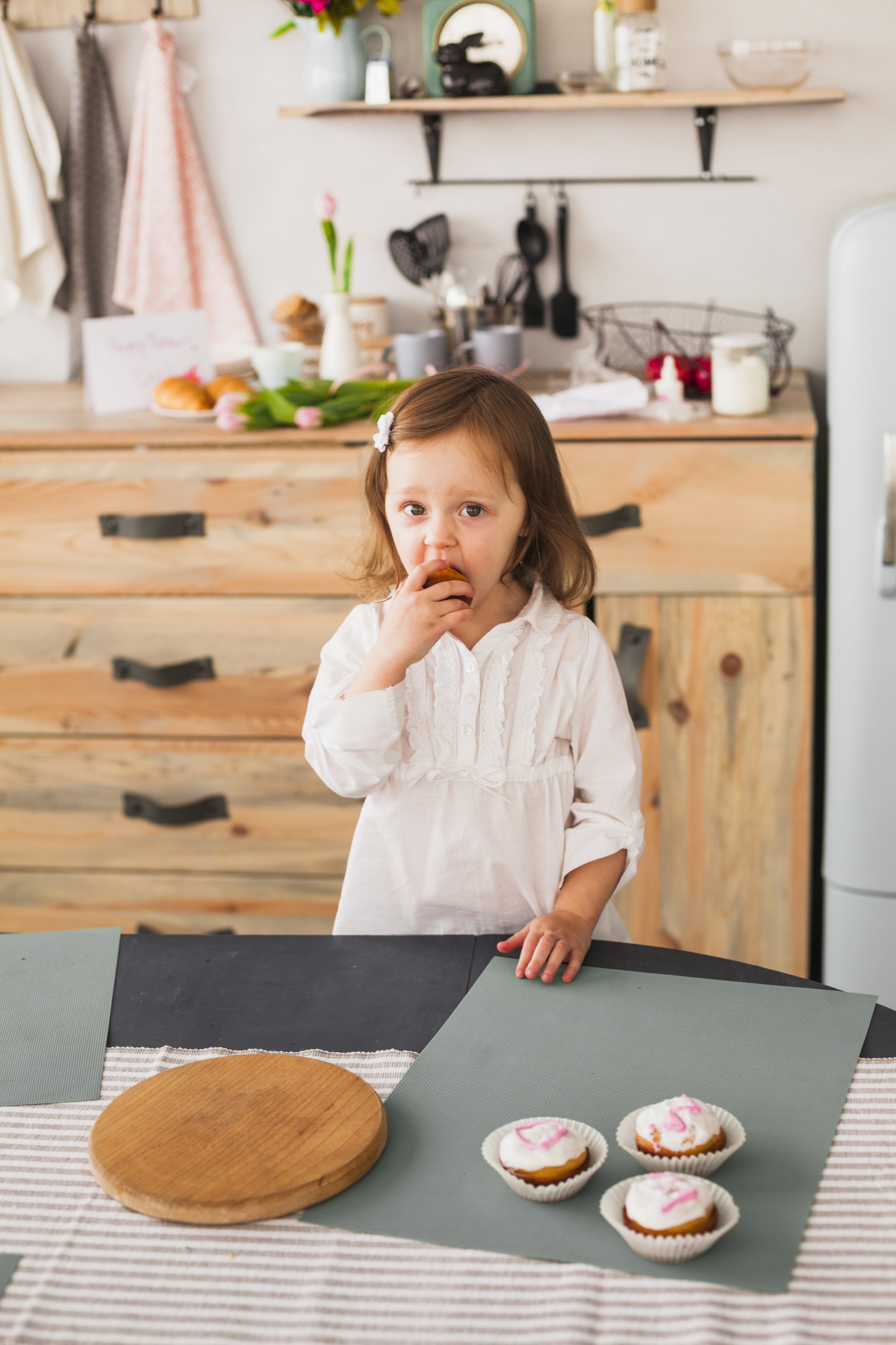 A young girl with brown hair wearing a white dress is standing at a table eating a cupcake. The table has three cupcakes with pink decorations, a round wooden cutting board, and green placemats. The kitchen background includes a wooden cabinet, shelves, and various kitchen items.
