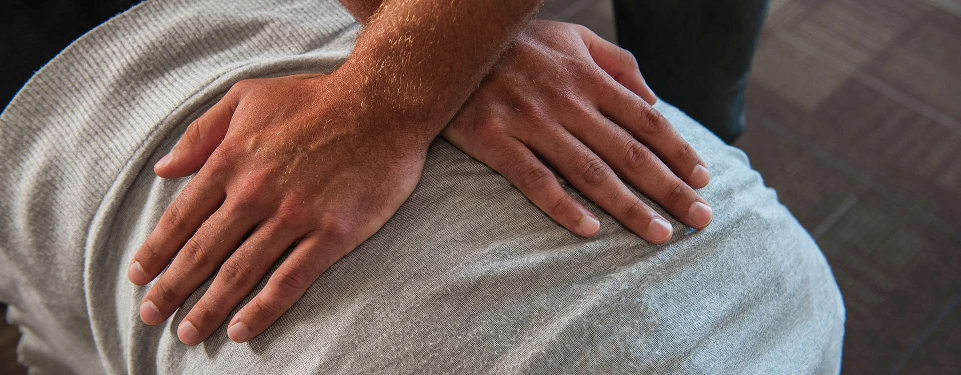 A person's hand resting on their knee, wearing a light gray, knit fabric clothing, with a dark wooden floor in the background.