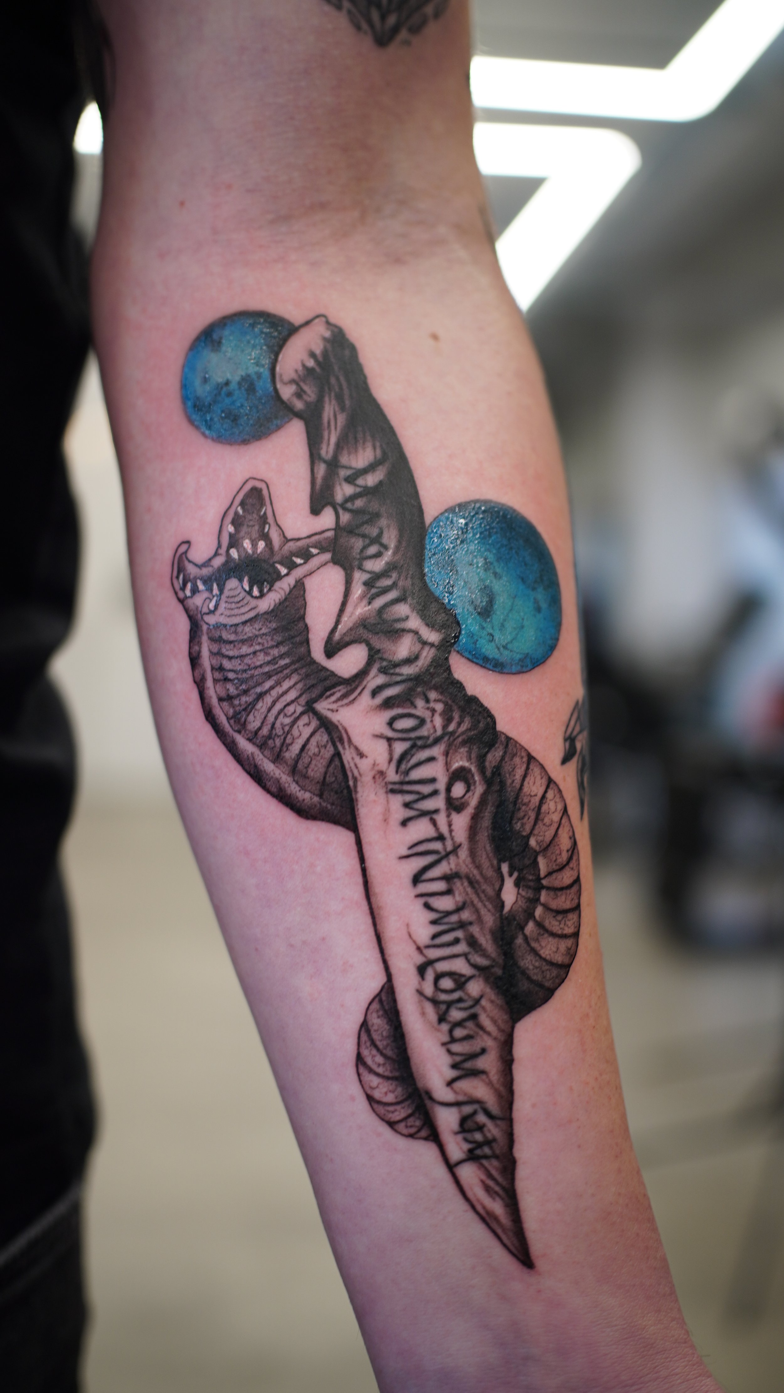 Tattoo of a snake wrapping around a sword with two planets and a quote on the forearm.