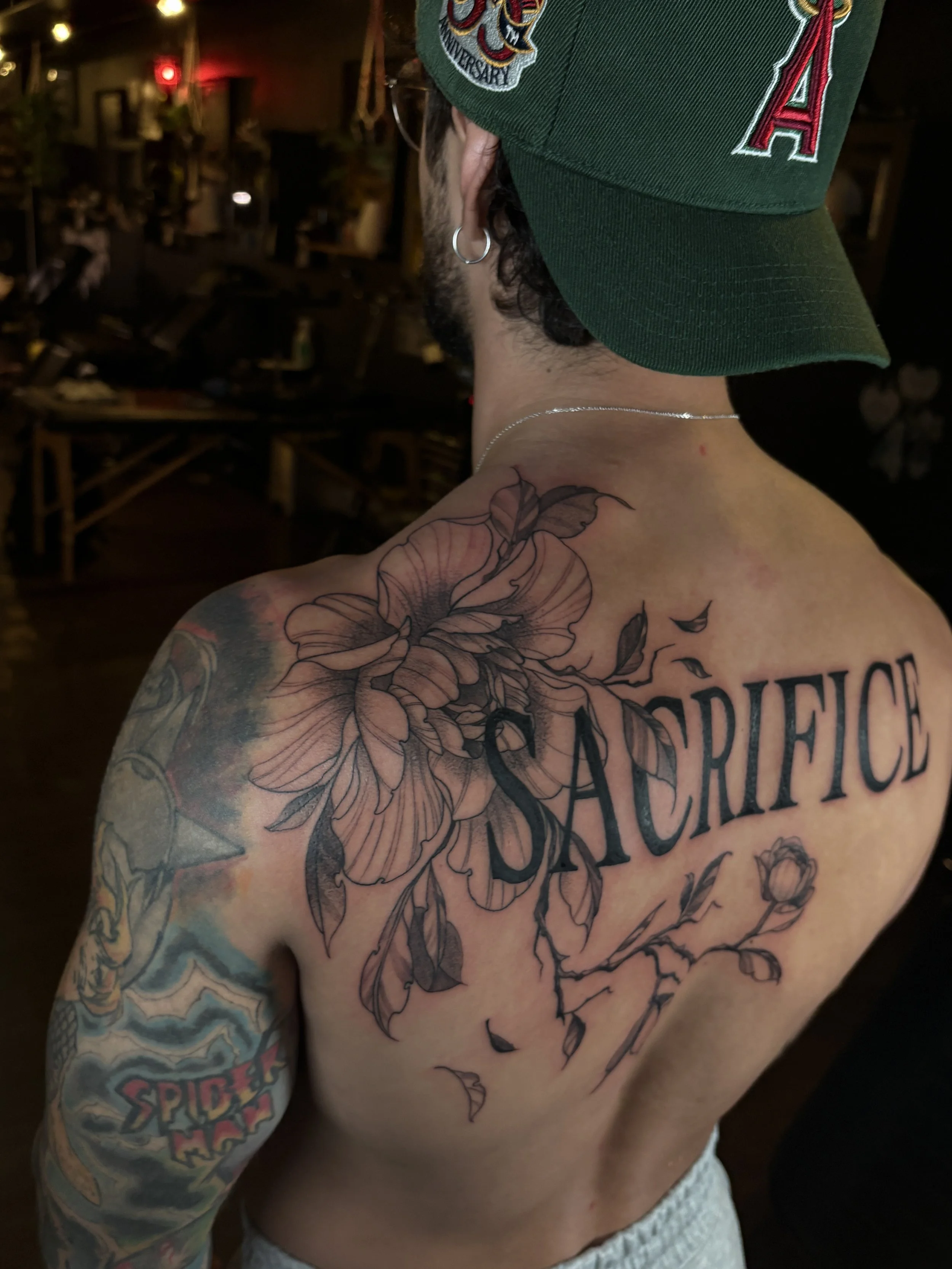A person with dark hair wearing a green baseball cap with patches and large earrings, displaying a large floral tattoo on their chest and a sleeve tattoo on their arm, in a dimly lit indoor setting.