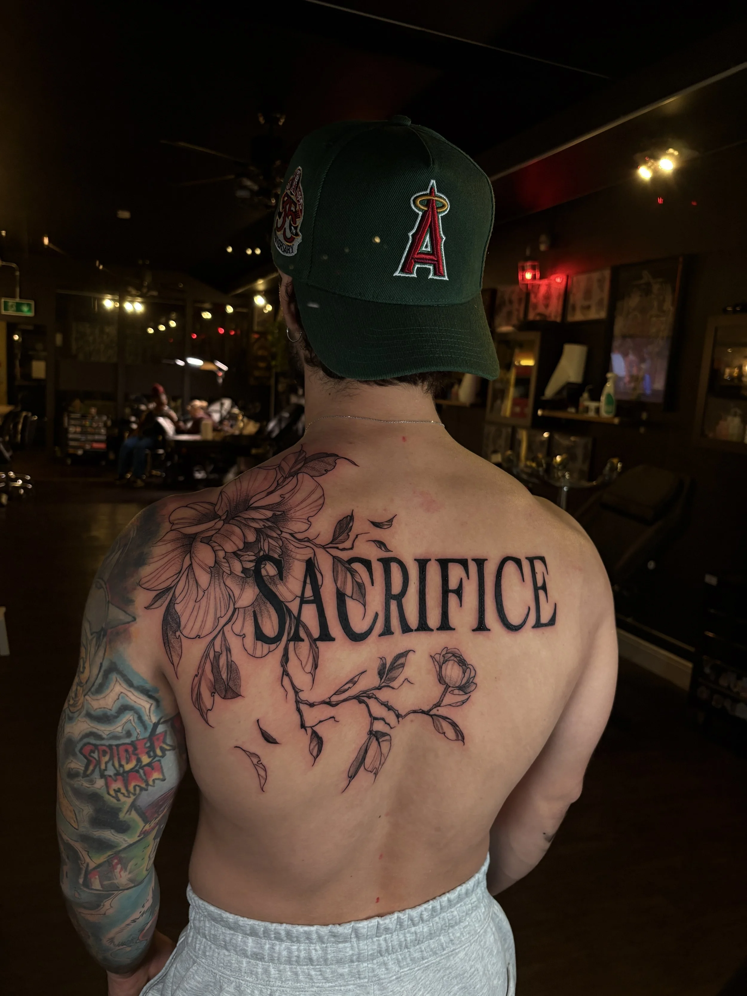 A person with tattoos on their back and arm, wearing a green Los Angeles Angels baseball cap, standing in a dimly lit room with a bar or restaurant setting.
