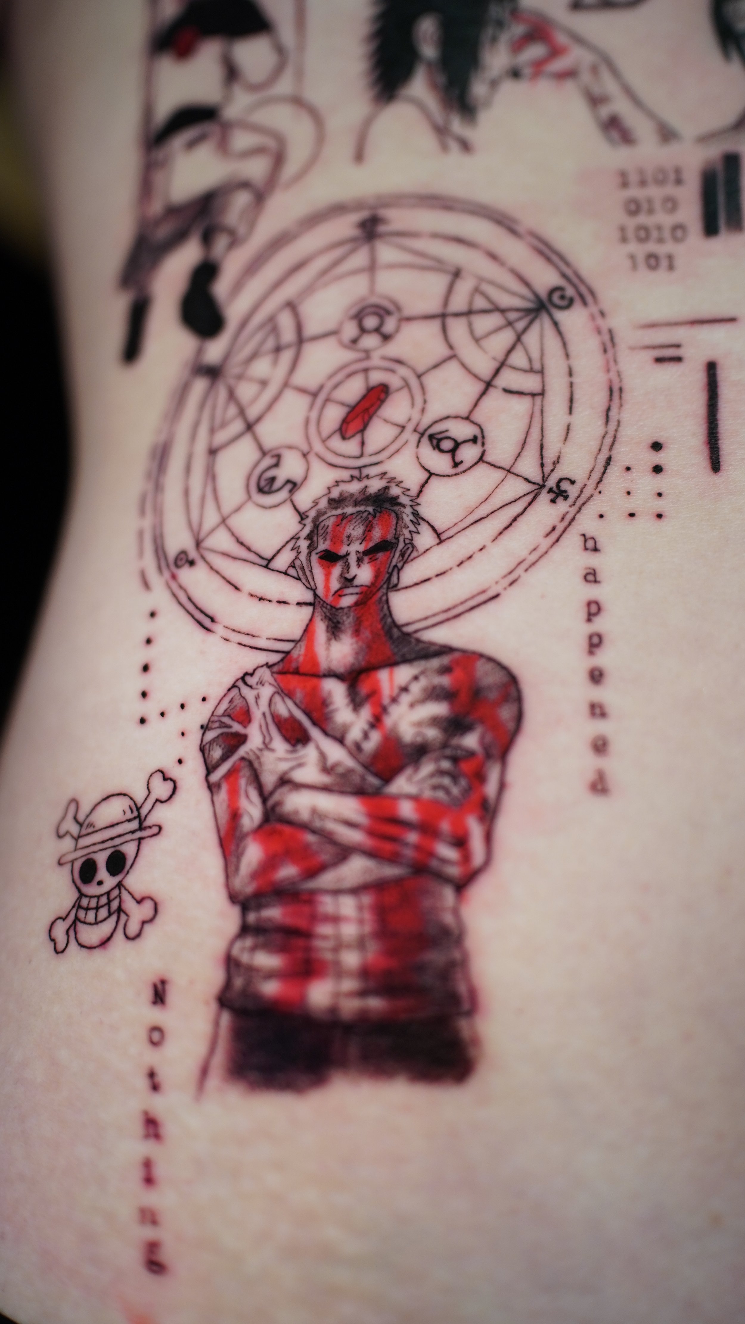 A tattoo featuring a character with cross-armed pose, short hair, and markings on the face and body, surrounded by geometric shapes, symbols, and tiny skulls. The tattoo includes red, black, and gray ink details.