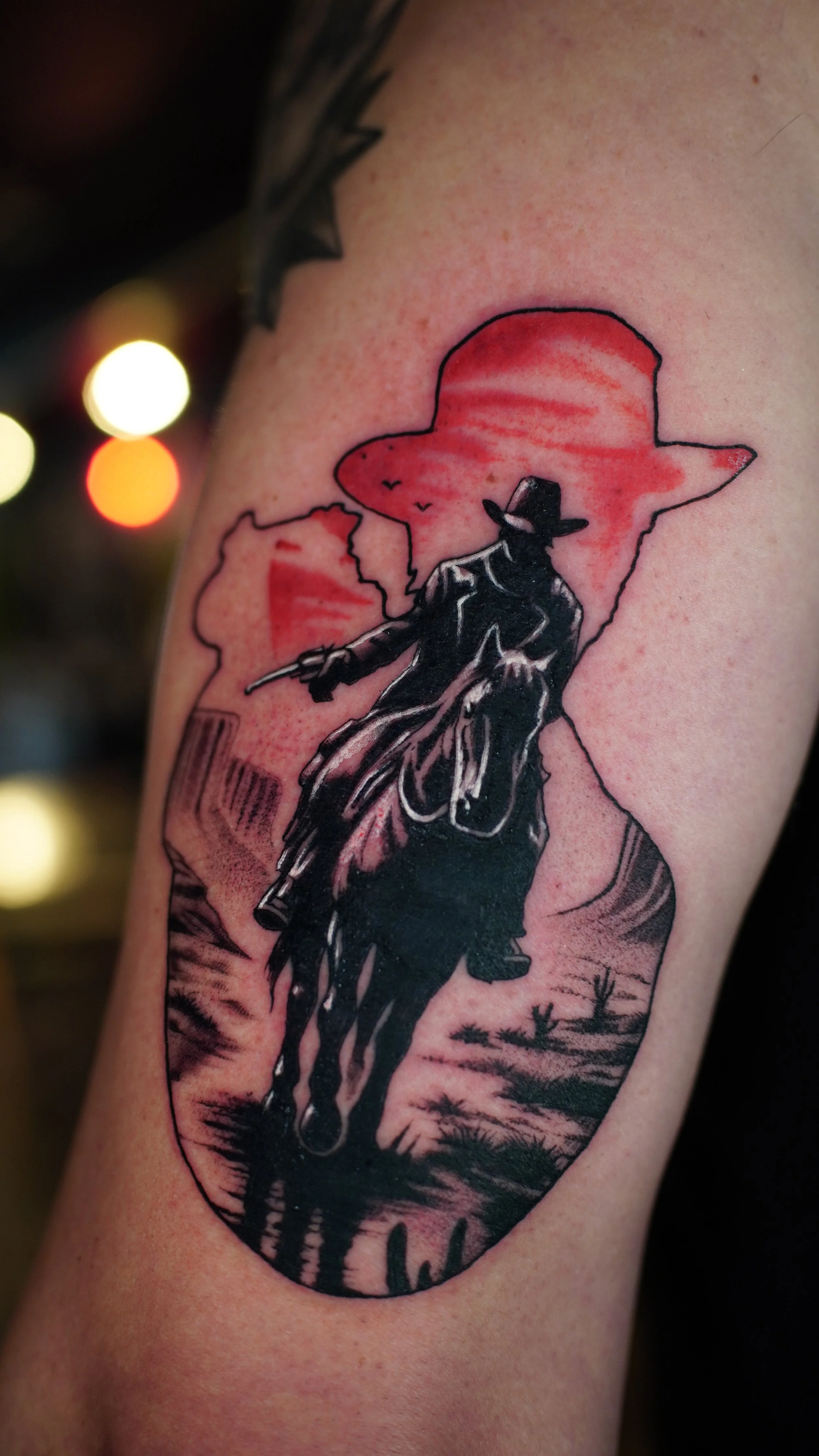 Tattoo of a cowboy riding a horse in front of a large red sunset with a star-shaped outline in the background.