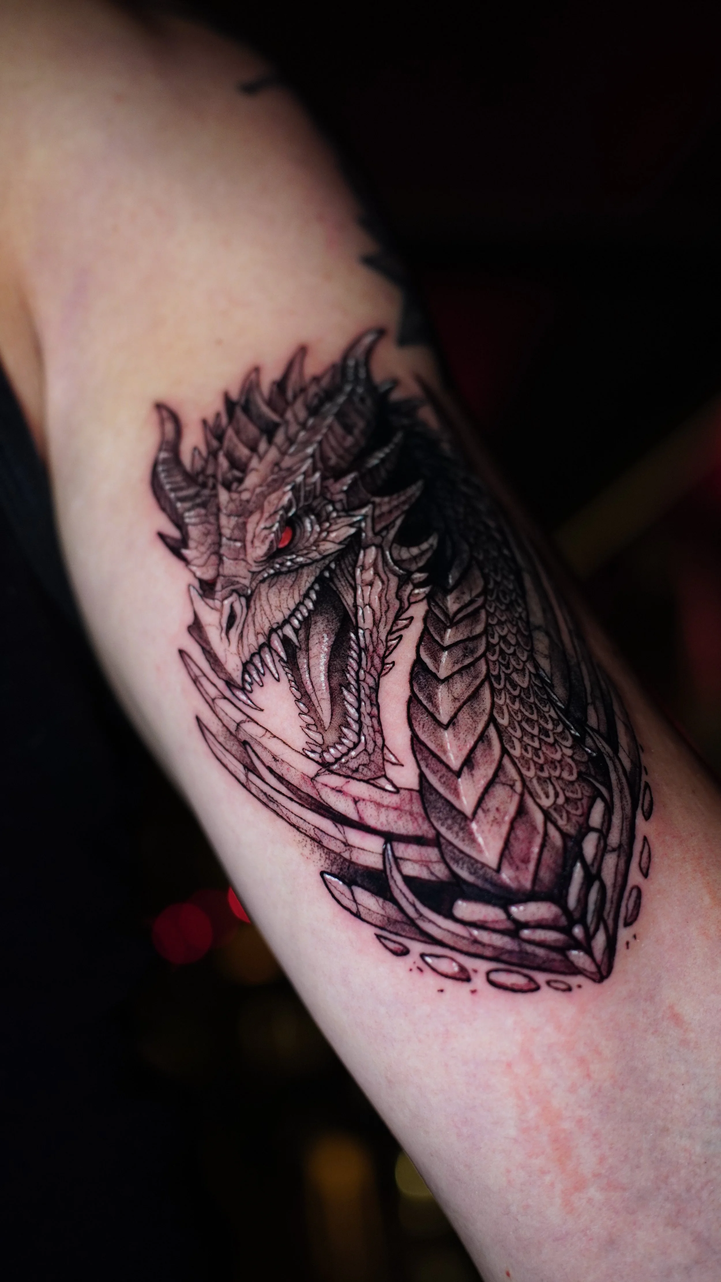 A black and gray dragon tattoo on a person's upper arm, featuring sharp scales, horns, and an open mouth showing teeth and tongue.