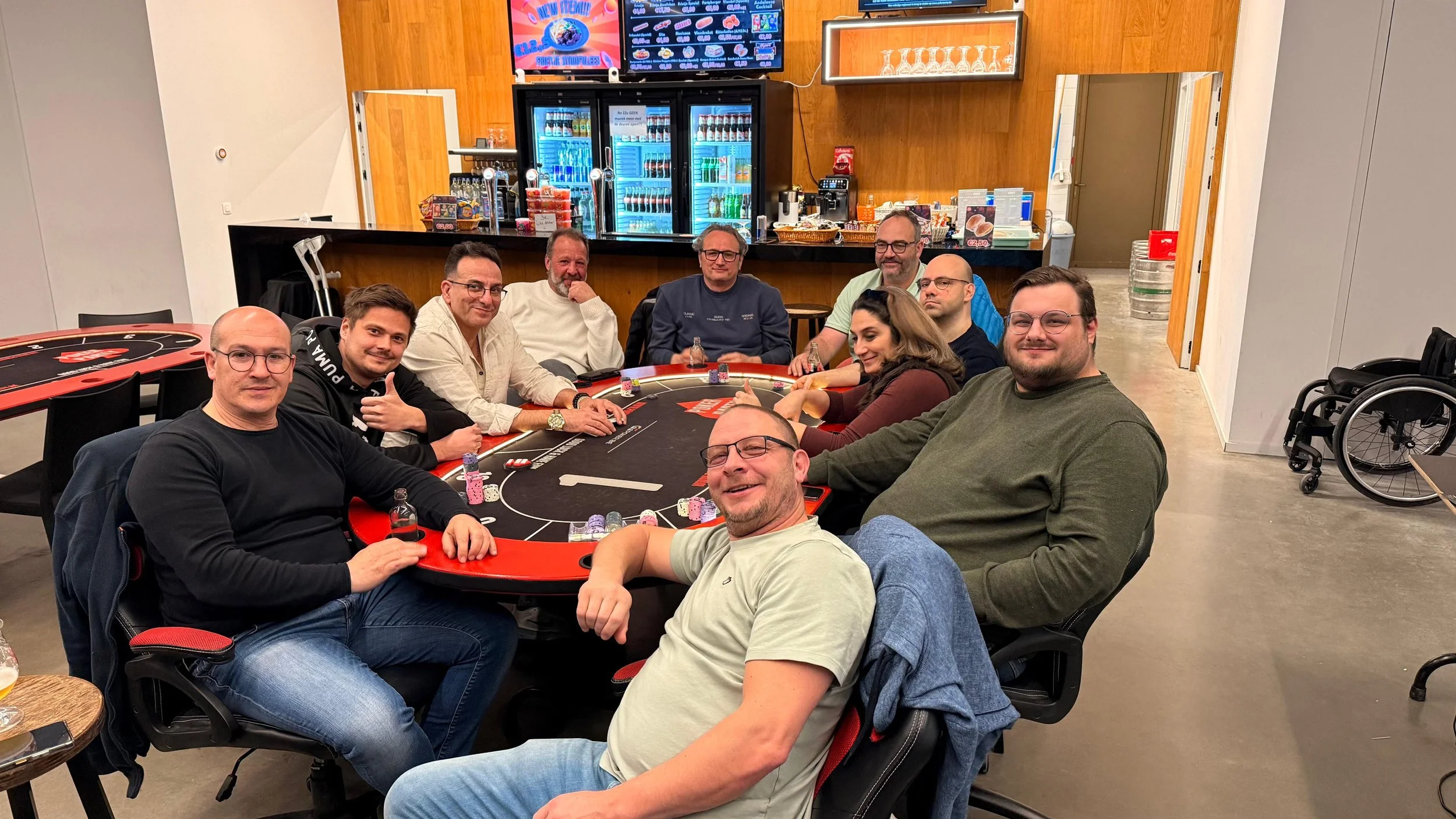 Een groep mensen die poker speelt aan een tafel in een casino of club, met een bar op de achtergrond en een rolstoel zichtbaar.