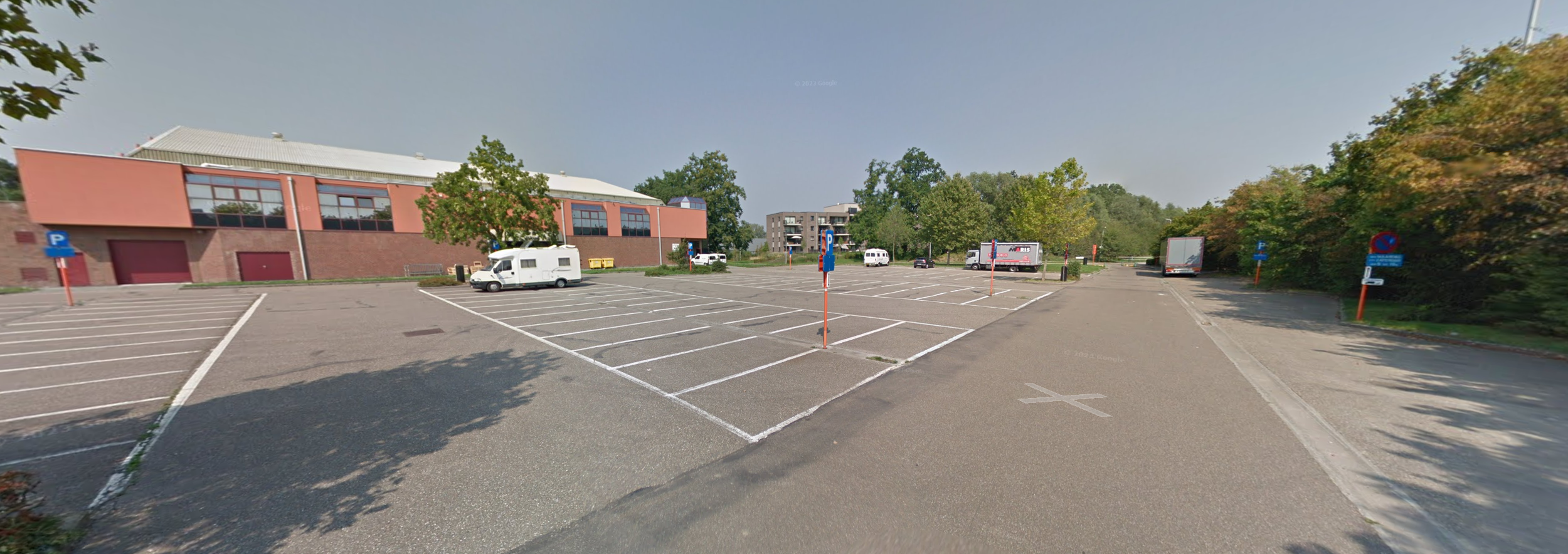 Een leeg parkeerterrein met enkele geparkeerde auto's en vrachtwagens, omgeven door bomen en een gebouw met grote ramen.