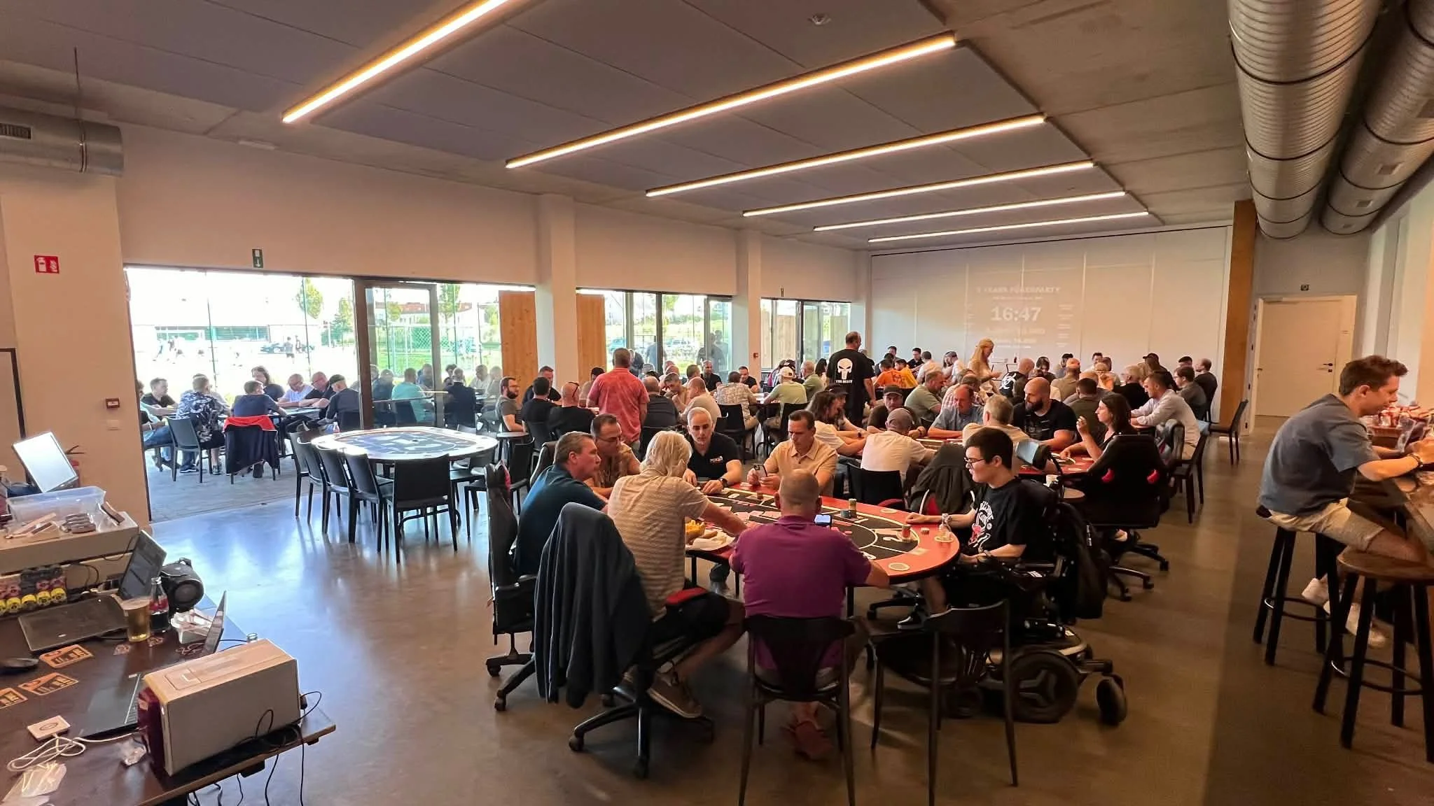 Grote zaal met veel mensen die pokertoernooi spelen aan tafel met pokerspellen, in de achtergrond grote ramen en een projectiescherm met tijdweergave.
