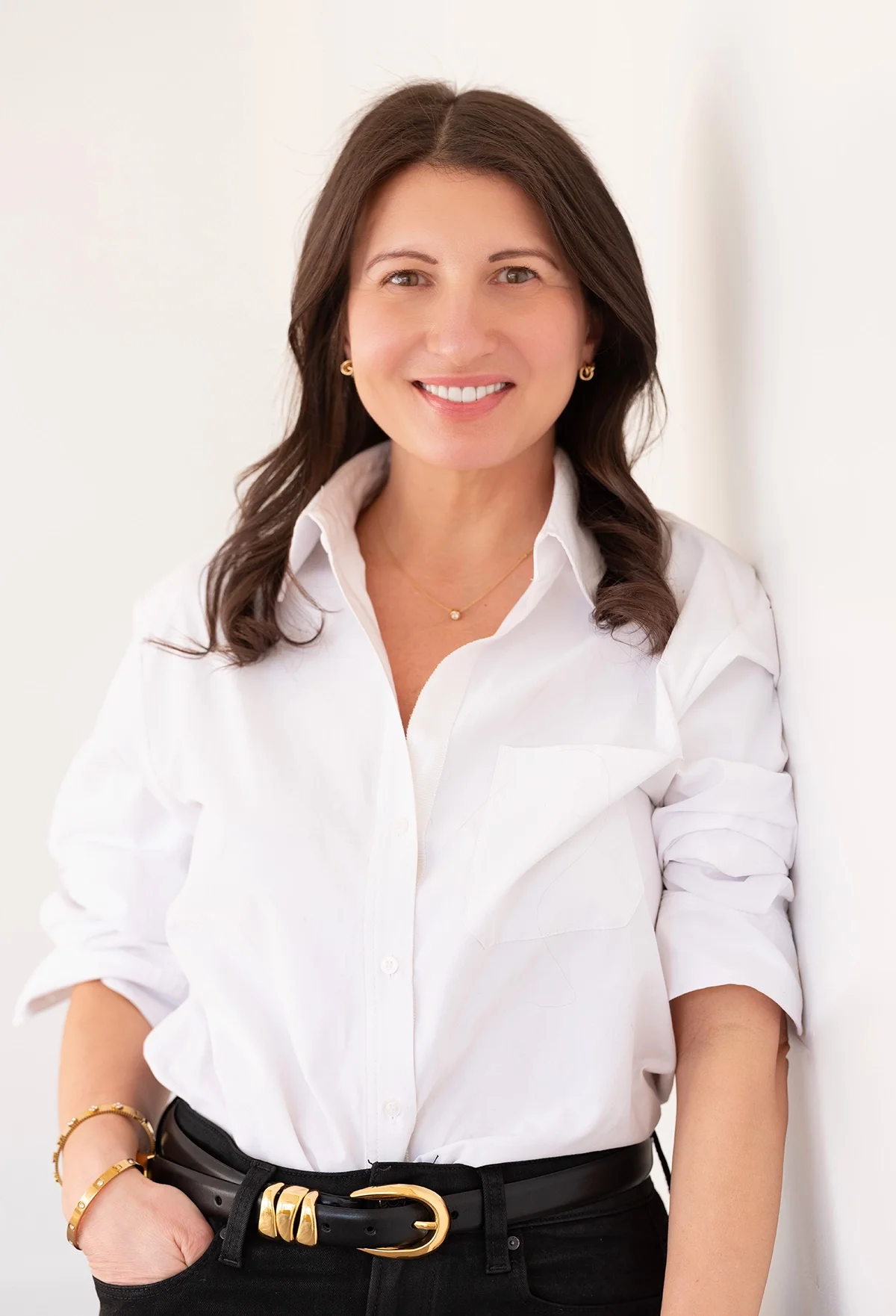 A woman with dark brown hair and light skin, smiling, wearing a white button-down shirt with rolled-up sleeves, black pants with a gold and black belt, gold jewelry, and standing against a light background.