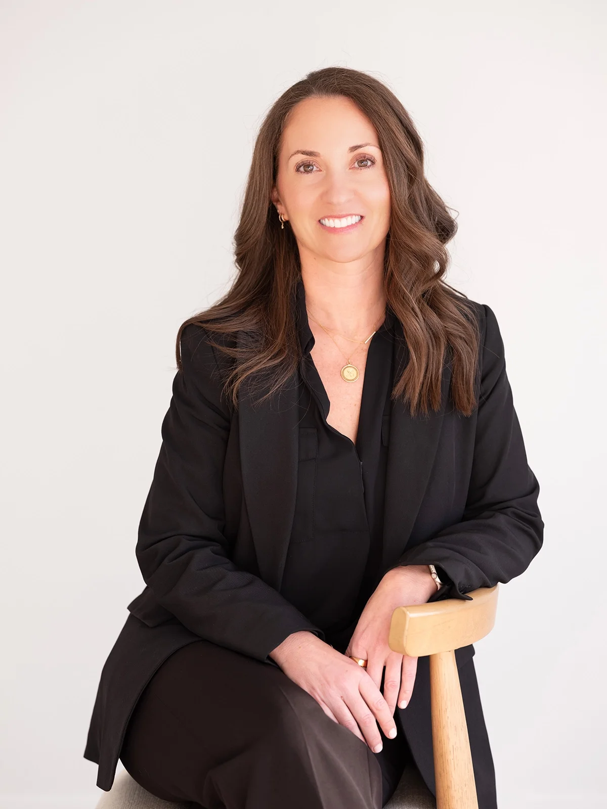 A woman with brown hair, wearing a black blazer and black shirt, sitting on a wooden stool against a white background.