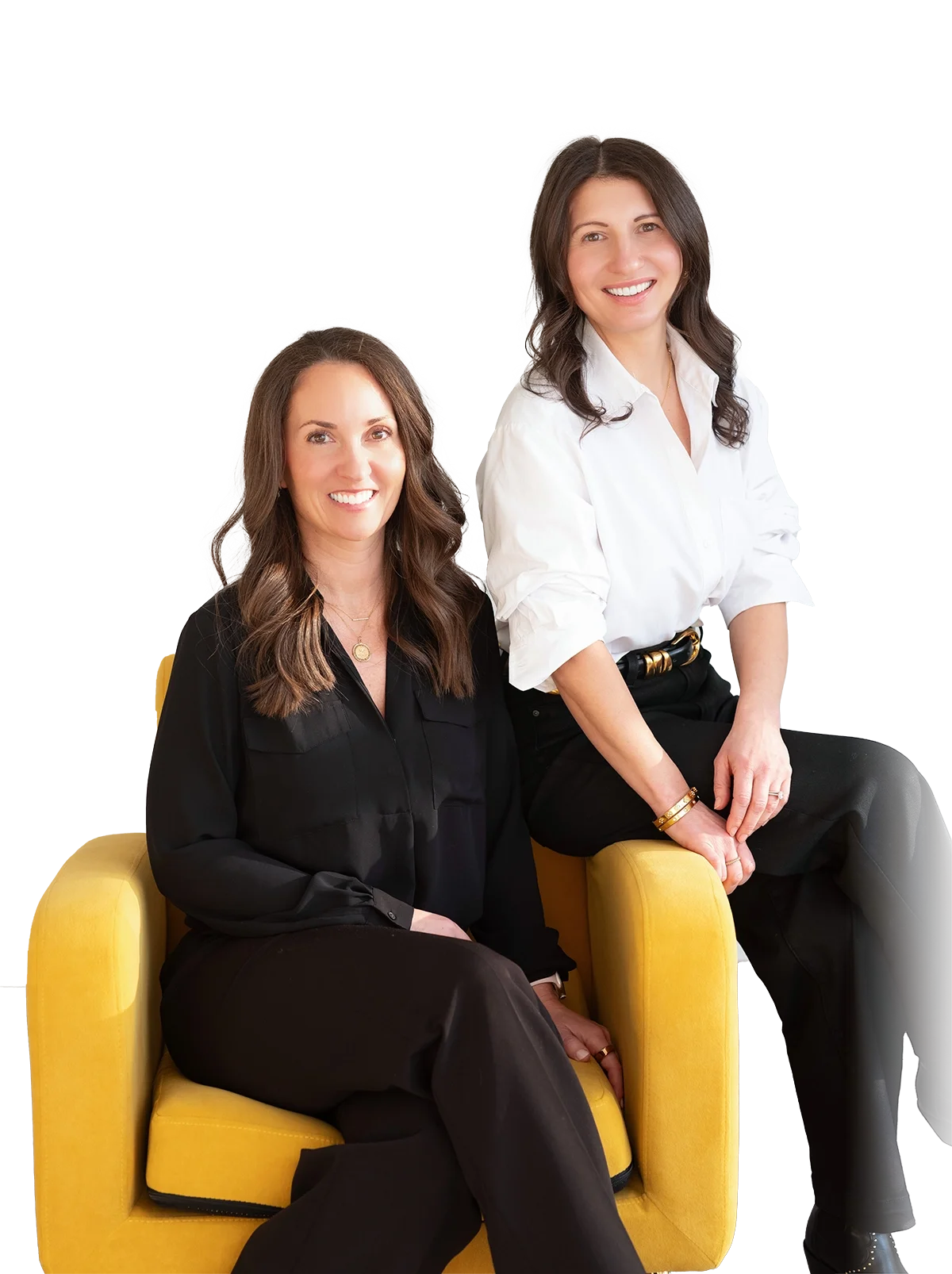 Two women with long brown hair smiling, sitting on a yellow couch, dressed in black and white shirts, against a plain white background.