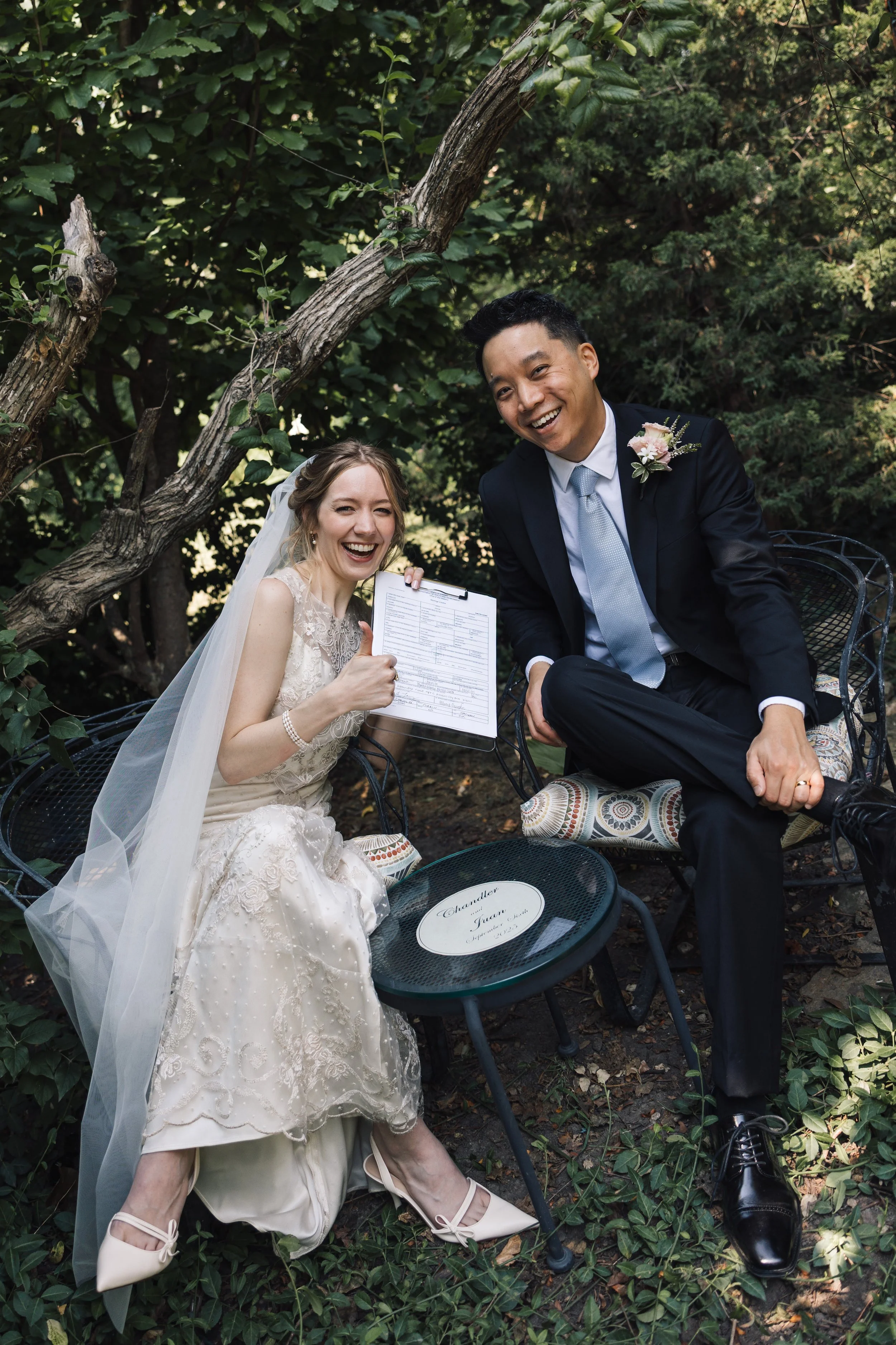 A bride and groom sitting outdoors among green trees and foliage, smiling and laughing. The bride is wearing a white lace wedding dress and veil, holding a clipboard, and her shoes are visible. The groom is wearing a dark suit with a white shirt and 