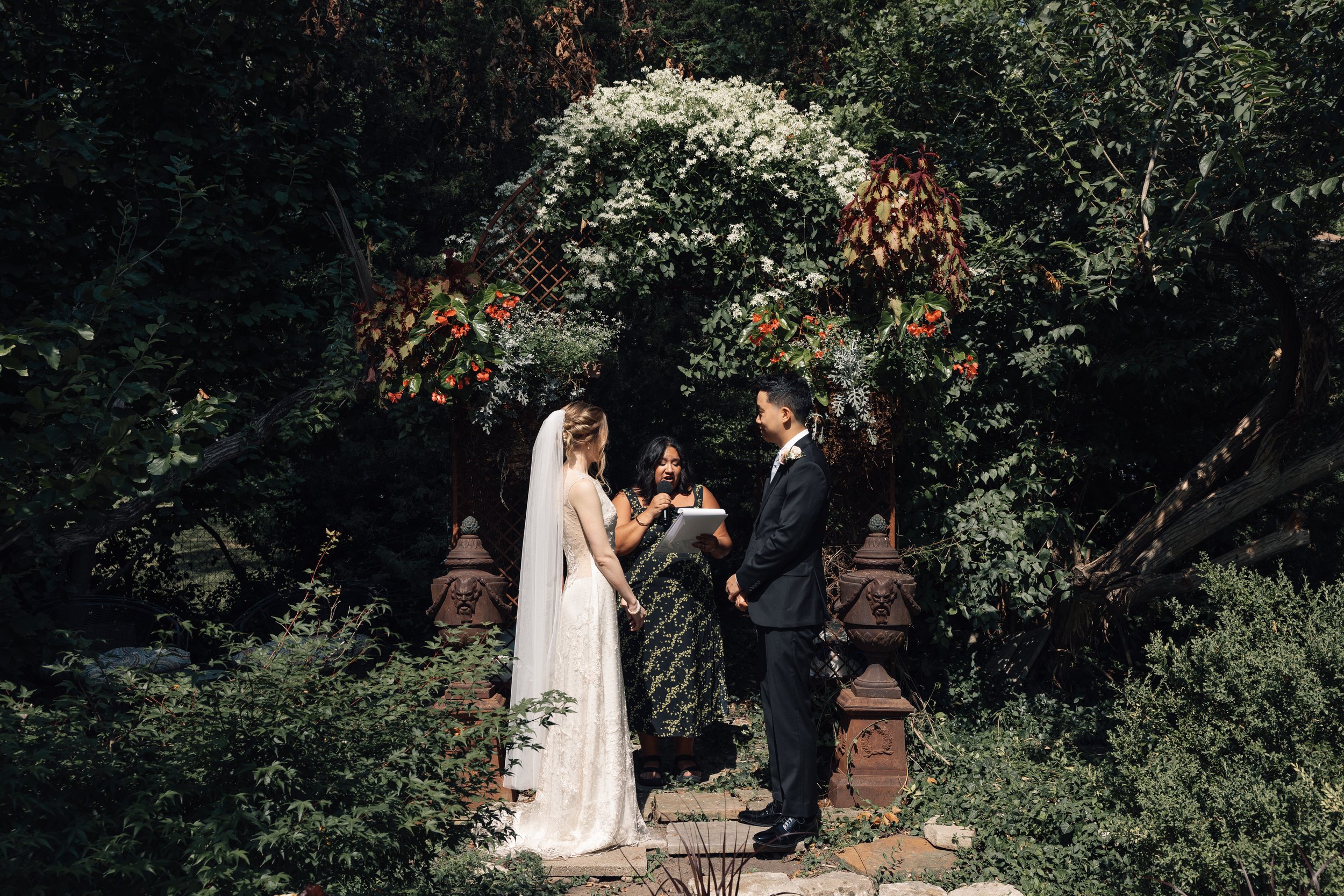A couple getting married outdoors under a floral arch, with a female officiant holding a microphone and a tablet, surrounded by lush greenery.