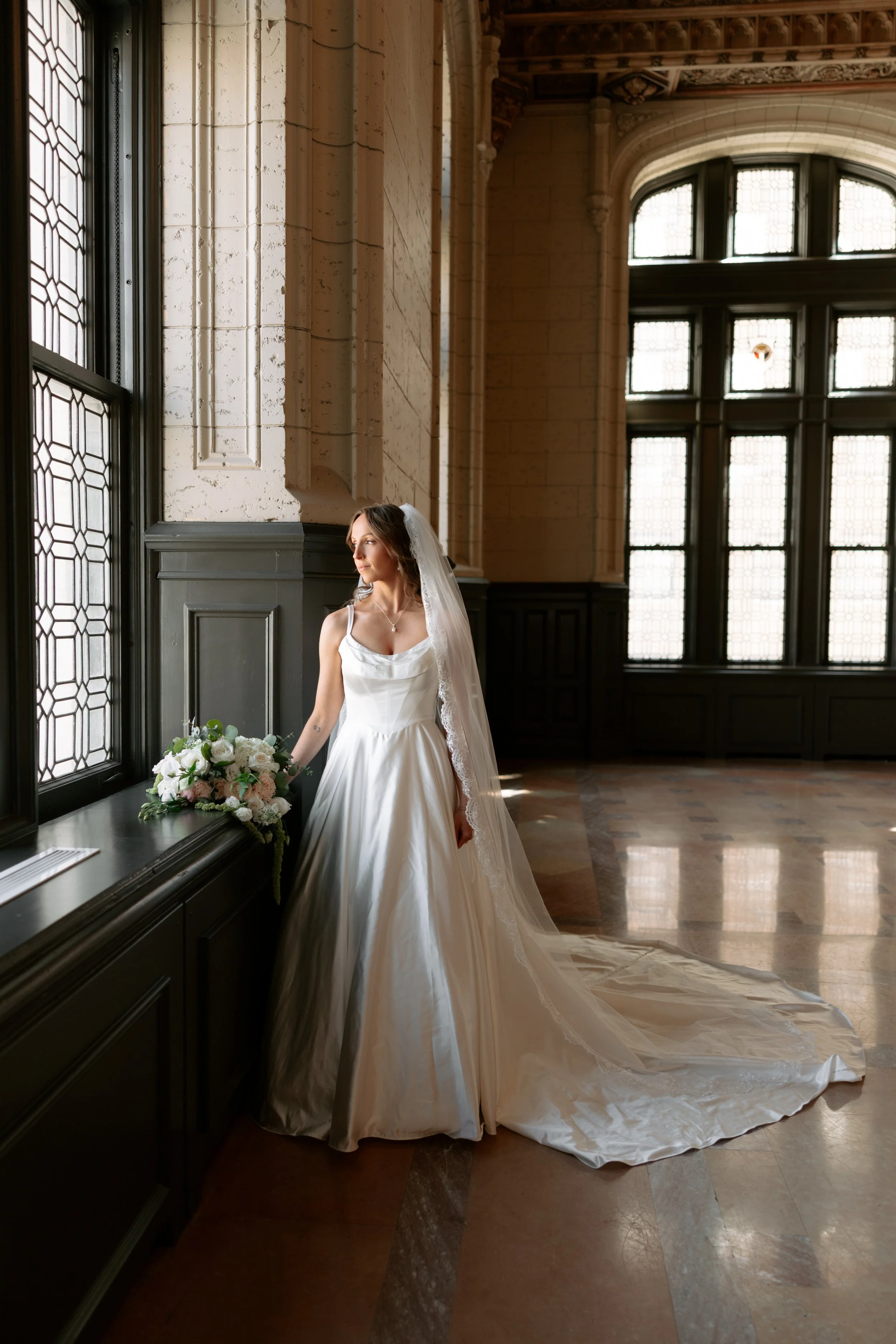 A bride in a white wedding dress and veil standing next to large windows in an elegant, historic building with intricate architectural details, looking out thoughtfully.