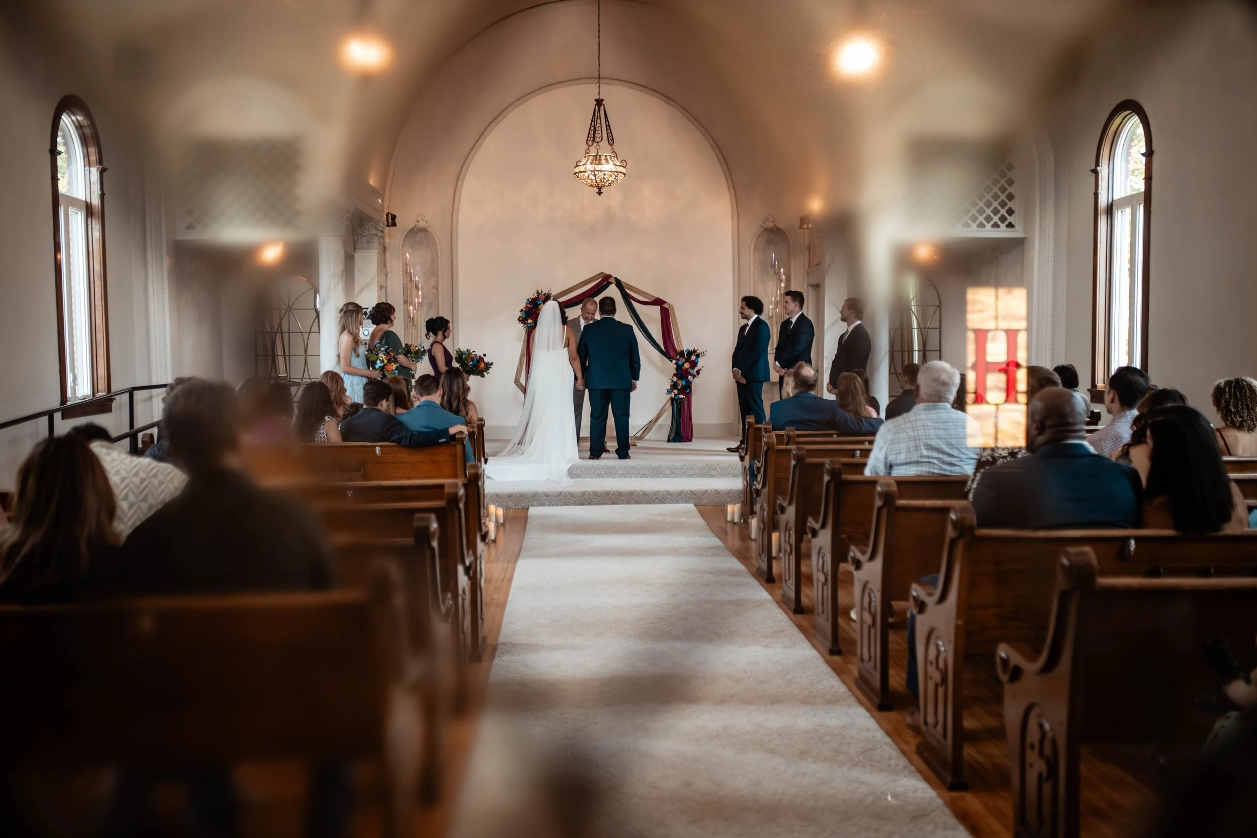 A wedding ceremony taking place inside a church with an arched ceiling, a chandelier, and stained glass windows. The bride and groom are standing at the altar with the officiant, surrounded by wedding party and guests seated in pews.