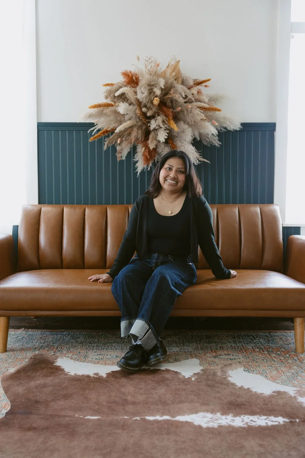A woman with dark hair and a black top sitting on a tan leather couch in a modern room with a large dried floral arrangement hanging on the wall behind her.