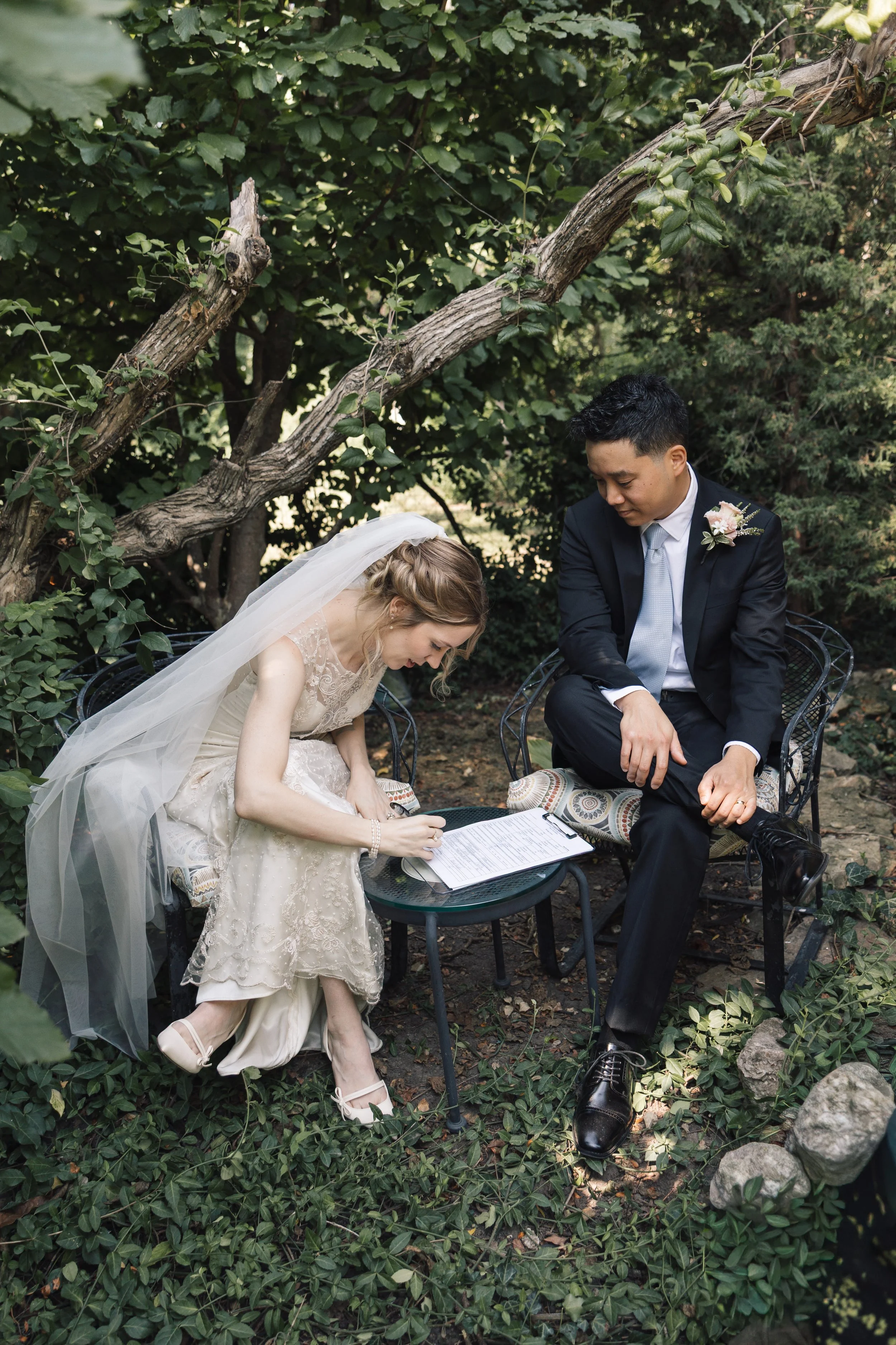 A bride and groom sitting outdoors on black metal chairs with leafy green surroundings. The bride is wearing a beige lace wedding dress and veil, writing on a document, while the groom, in a black suit, white shirt, and light gray tie with a floral b