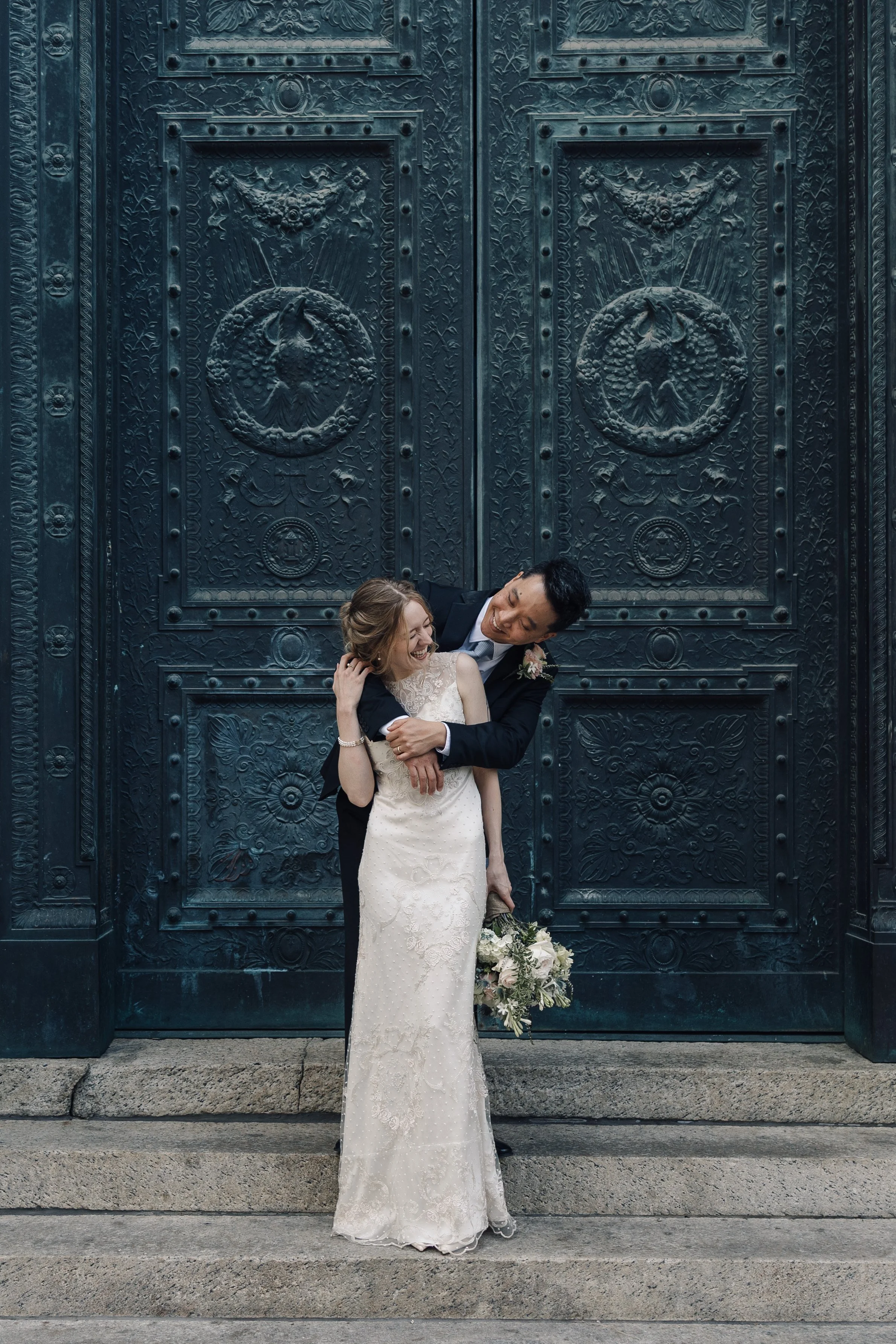 A newlywed couple in wedding attire standing on steps in front of an ornate, large dark metal door with intricate designs and patterns. The bride is wearing a white lace wedding dress and holding a bouquet, and the groom is in a black suit with a floral boutonniere, embracing and smiling.
