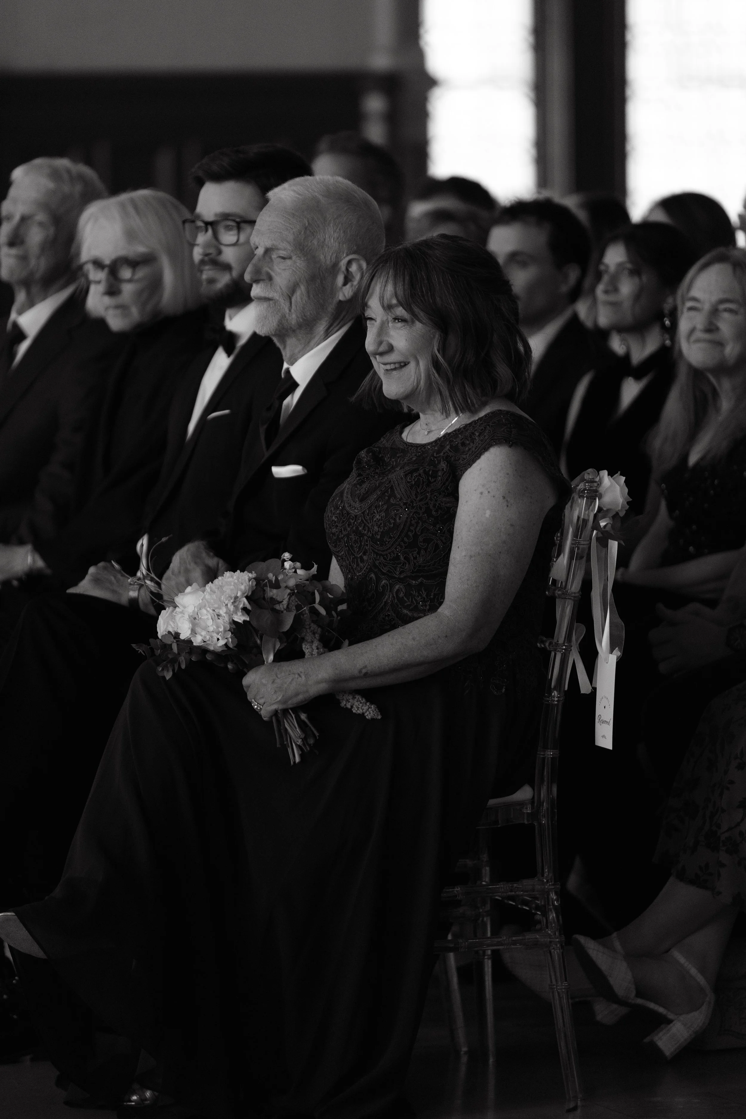 Black and white photo of a group of people dressed in formal attire, seated and attending an event. The focus is on a woman with shoulder-length hair smiling, holding a bouquet, and sitting next to an elderly man with a beard, both dressed in suits. 